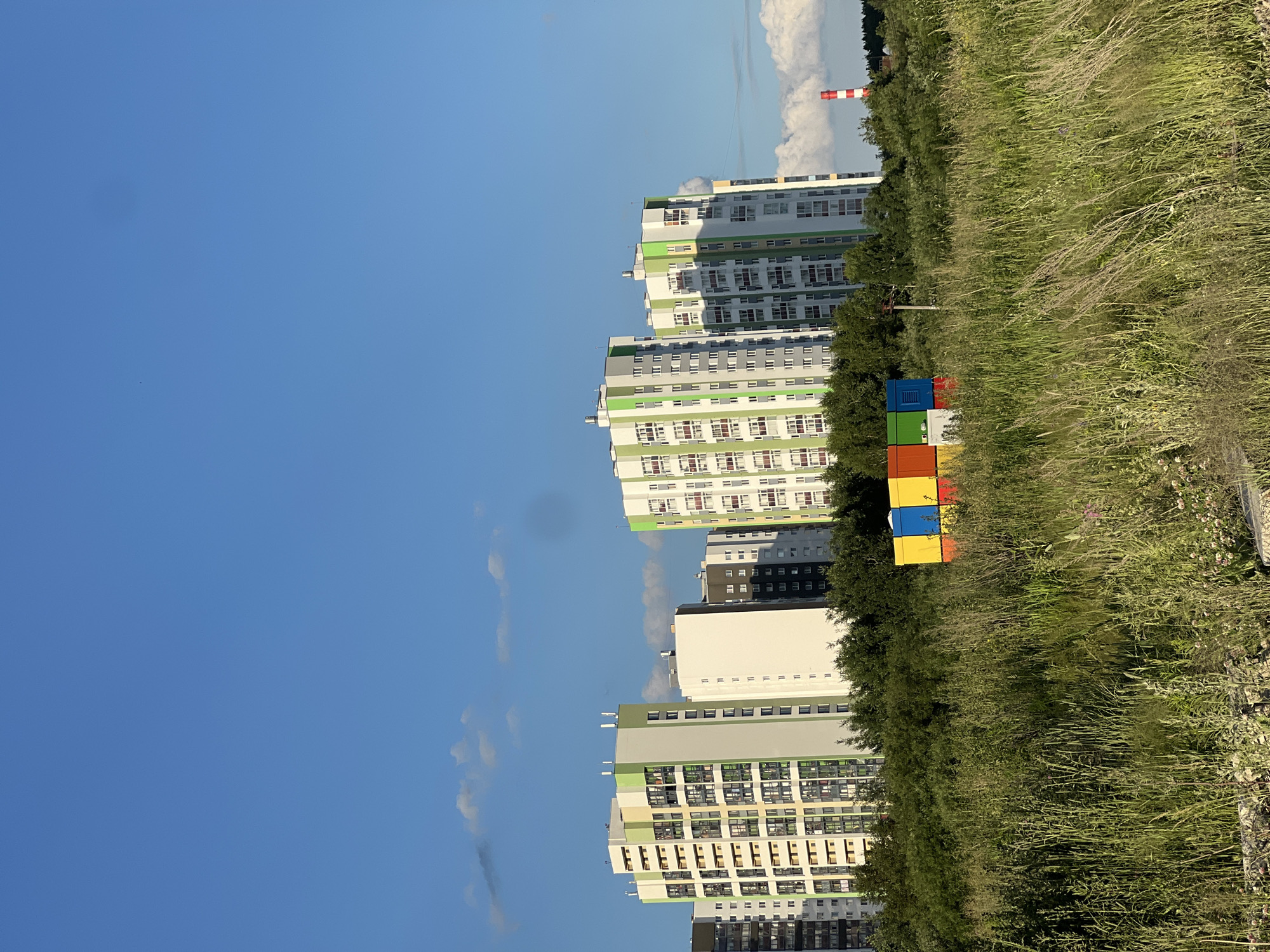 The image captures a vibrant urban landscape with a clear blue sky dotted with sparse white clouds. In the foreground, a field of tall, green grass stretches out, with some smaller pink flowers visible. In the middle ground, a cluster of brightly colored modular boxes—red, blue, yellow, and green—are stacked against a backdrop of dense green trees.
Behind the trees, a row of modern, multi-story apartment buildings stands tall. These buildings feature a striking architectural design with alternating sections of white, light green, and light yellow facades, accented by numerous windows. The buildings are arranged in a slight arc, suggesting a planned residential development.
Further in the background, to the right, a tall, slender industrial chimney with red and white stripes is visible, hinting at the presence of industrial activity in the vicinity. The overall scene suggests a developing urban area where modern residential structures are being integrated with natural elements and potentially some industrial infrastructure. The image appears to be taken during daylight hours, with the sun casting gentle shadows.