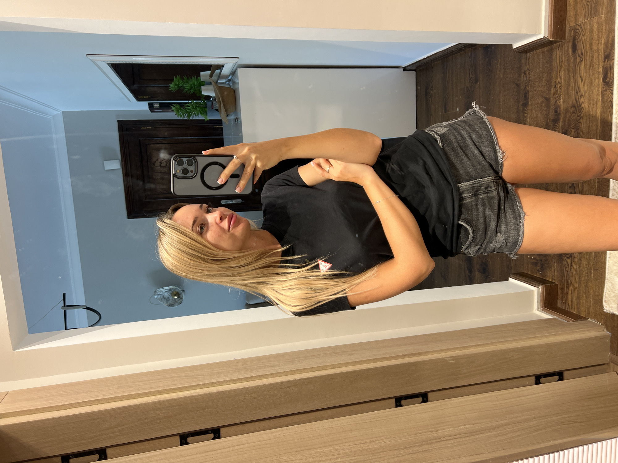 A young, blonde woman with fair skin and light makeup takes a selfie in a hallway. She is wearing a black Guess t-shirt and distressed denim shorts. She is holding up her phone to take the picture and her right arm is resting on her left arm, which is bent at the elbow. There is a mirror behind her that reflects the hallway, a dark wooden door, a white cabinet with plants, and a straw hat. The floor is made of dark wood planks, and there is a white baseboard along the wall. The location is a residential hallway in Sharm El Sheikh, Egypt.