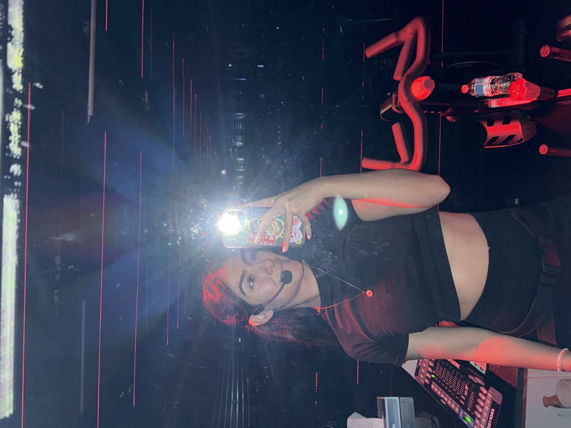 A woman is taking a selfie. She is wearing a headset microphone and a dark crop top. She is standing in front of a wall with vertical red and white light bars. There is a stationary bike and a light control board in the frame. The location is a studio in Puebla, Mexico.