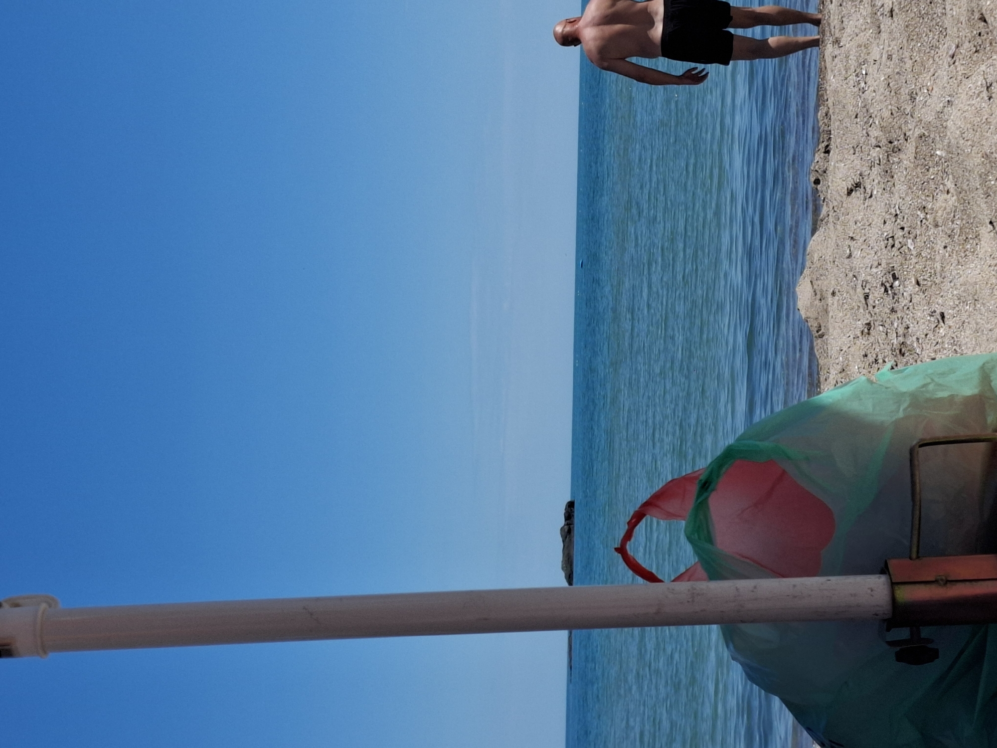 A person stands near the edge of the ocean on a sandy beach. The person is bald and wearing black swim trunks. The ocean is blue and calm, with a few rocks visible in the distance. In the foreground, a white pole and a green and pink bag are present. The sky is clear and blue.