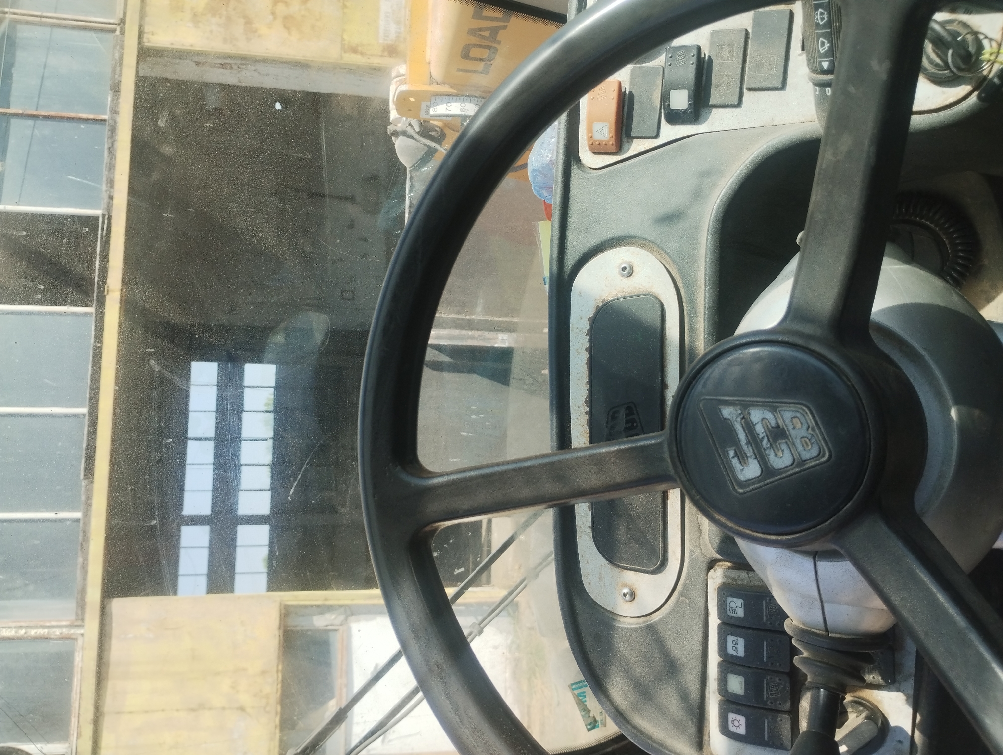 The image shows the interior of a construction vehicle, likely a loader, as viewed from the driver's seat. The steering wheel is black and prominently features the JCB logo. To the left of the steering wheel, a row of several toggle switches is visible, some with illuminated icons indicating functions like headlights. To the right and behind the steering wheel, various buttons and a gauge are present. The dashboard is dusty, suggesting the vehicle is used in an outdoor or construction environment. Through the dirty windshield, a building with large windows and a yellow structure with the word "LOAD" visible can be seen. The overall scene conveys a sense of industrial or agricultural activity. The location is Sredets, Bulgaria.