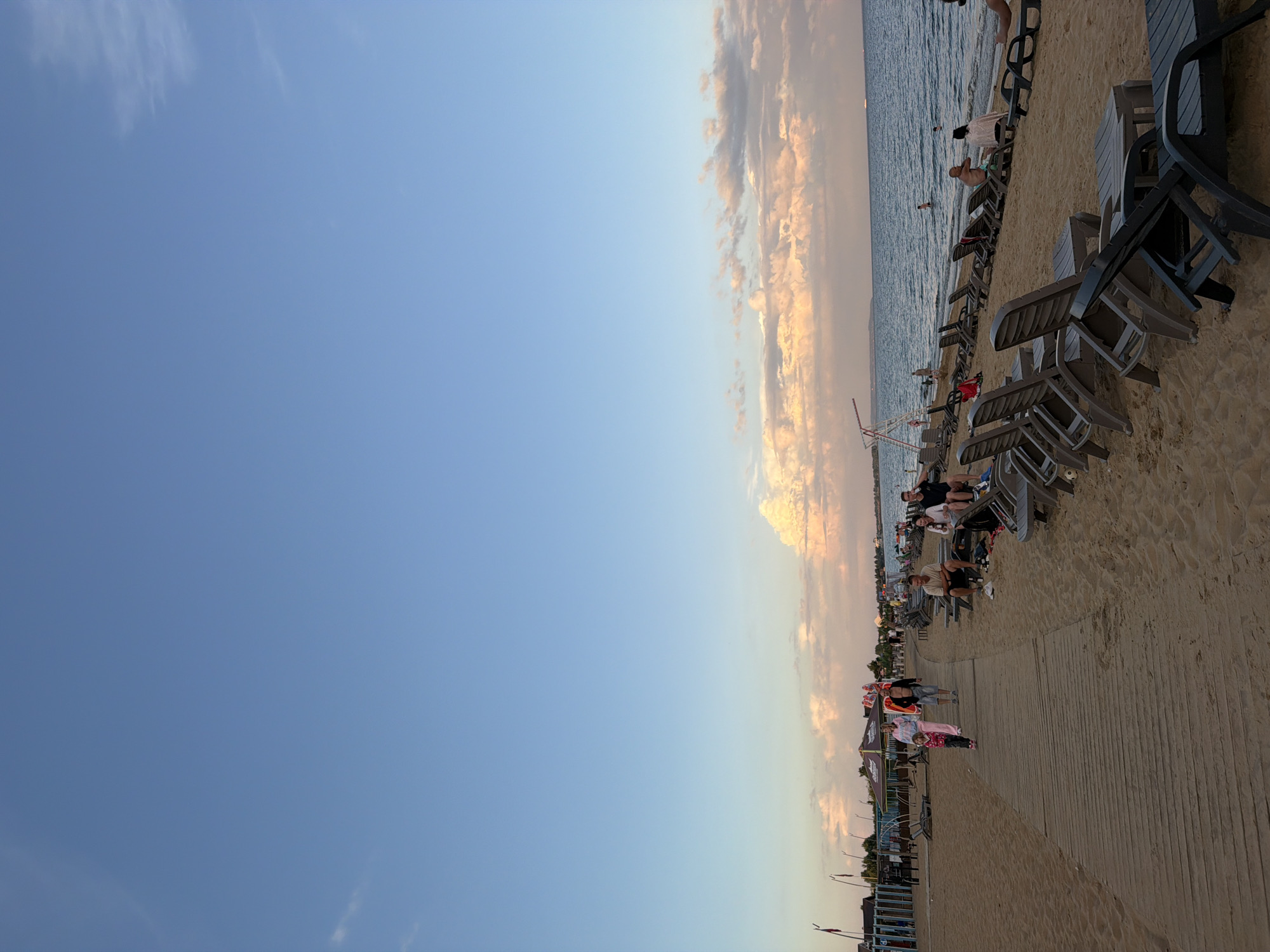 This image depicts a beach scene in Laishevo, Russia, during what appears to be late afternoon or early evening. The sky is a clear blue with some scattered clouds tinged with pink and orange from the setting sun. The beach is sandy, with a wooden boardwalk running through it. Rows of empty lounge chairs are set up on the sand, facing the water.
Several people are visible in the image. A family, including two adults and a young child, are walking along the boardwalk. Further down the beach, a group of people are seated on lounge chairs, some interacting, while others appear to be relaxing or observing the water. A few individuals are in the shallow water near the shore.
In the background, there are some structures that suggest amenities such as kiosks or changing rooms, along with some flags and umbrellas. The sea is calm, with a few distant boats visible on the horizon. The overall atmosphere is one of leisure and relaxation.