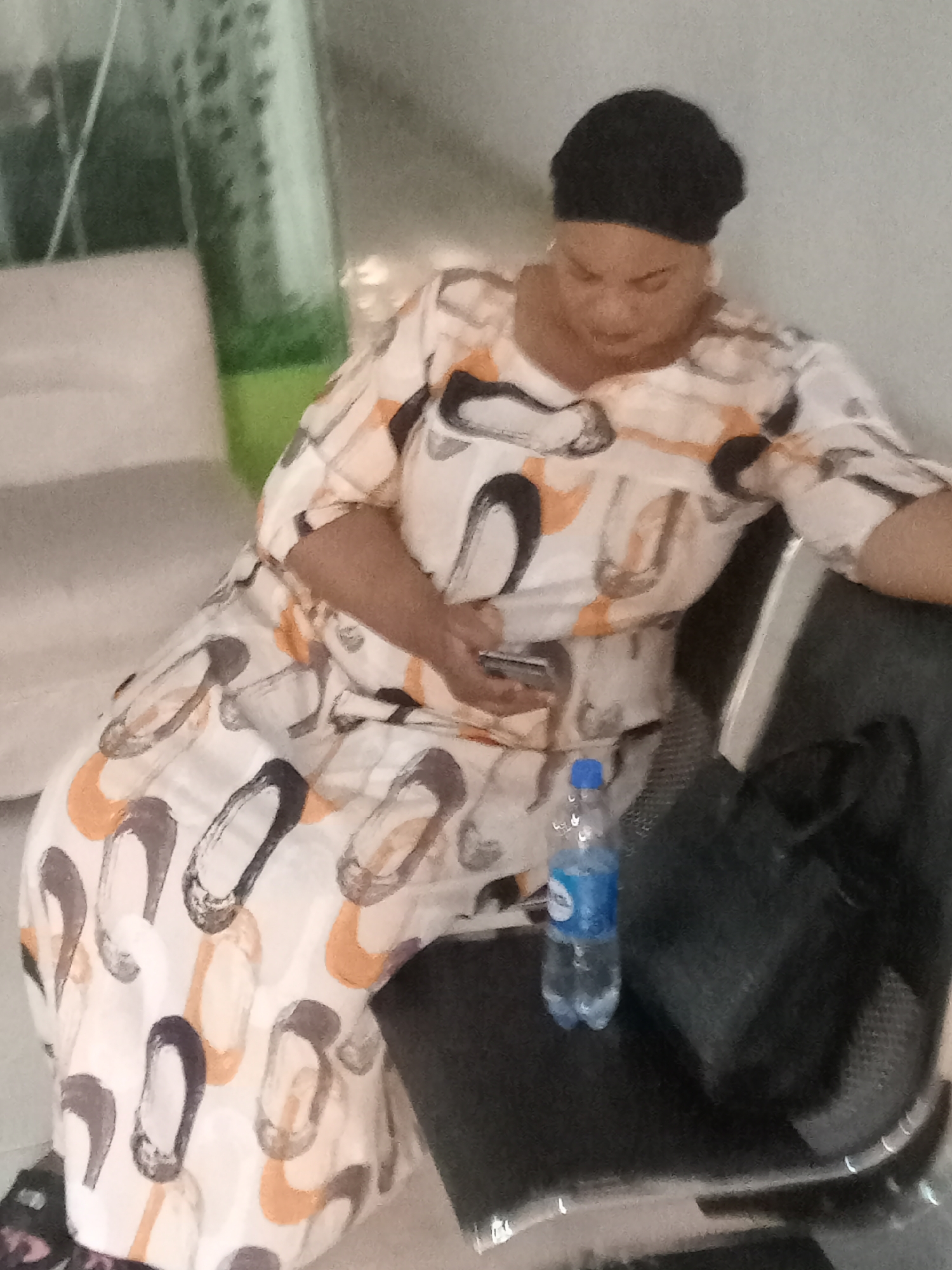 In Gwarinpa, Nigeria, a woman wearing a patterned dress and black head covering sits in a chair. She is holding a smartphone. A water bottle and a black handbag are on the seat next to her. The background includes a blurred glimpse of seating and a green object behind a partially transparent surface.