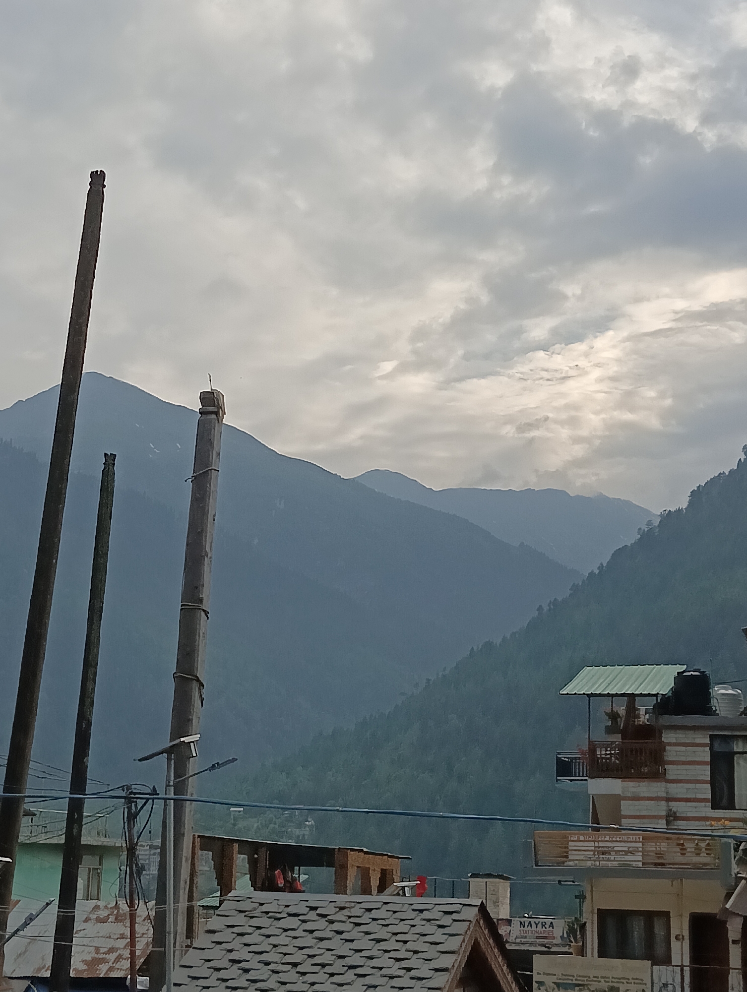 This image captures a serene mountainous landscape with a hint of a village settlement. In the foreground, three tall wooden poles stand vertically, silhouetted against a dramatic cloudy sky. The sky itself is filled with a mixture of light and dark grey clouds, suggesting either an overcast day or the approach of rain.
Behind the poles, majestic mountains rise in layers, their peaks fading into the hazy distance. The slopes of the mountains are densely covered with dark green pine trees, creating a lush and textured backdrop. Patches of snow are visible on some of the higher mountain ridges, hinting at a cooler climate.
At the base of the mountains, a glimpse of a village is visible. Several buildings with tiled roofs and multi-story structures with balconies can be seen. One building features a sign that reads "NAYRA STATIONARIES," indicating a commercial establishment. Another building has a green roof and a water tank on top. Power lines and streetlights are also present, suggesting a populated area.
The overall mood of the image is tranquil and slightly dramatic due to the cloudy sky and the imposing presence of the mountains. The time of day appears to be late afternoon or early evening, given the soft light and the diffused sky. The location is clearly Vashisht, India, as indicated by the name of the stationery shop. There is no discernible activity or event taking place; the scene is a static, picturesque view.