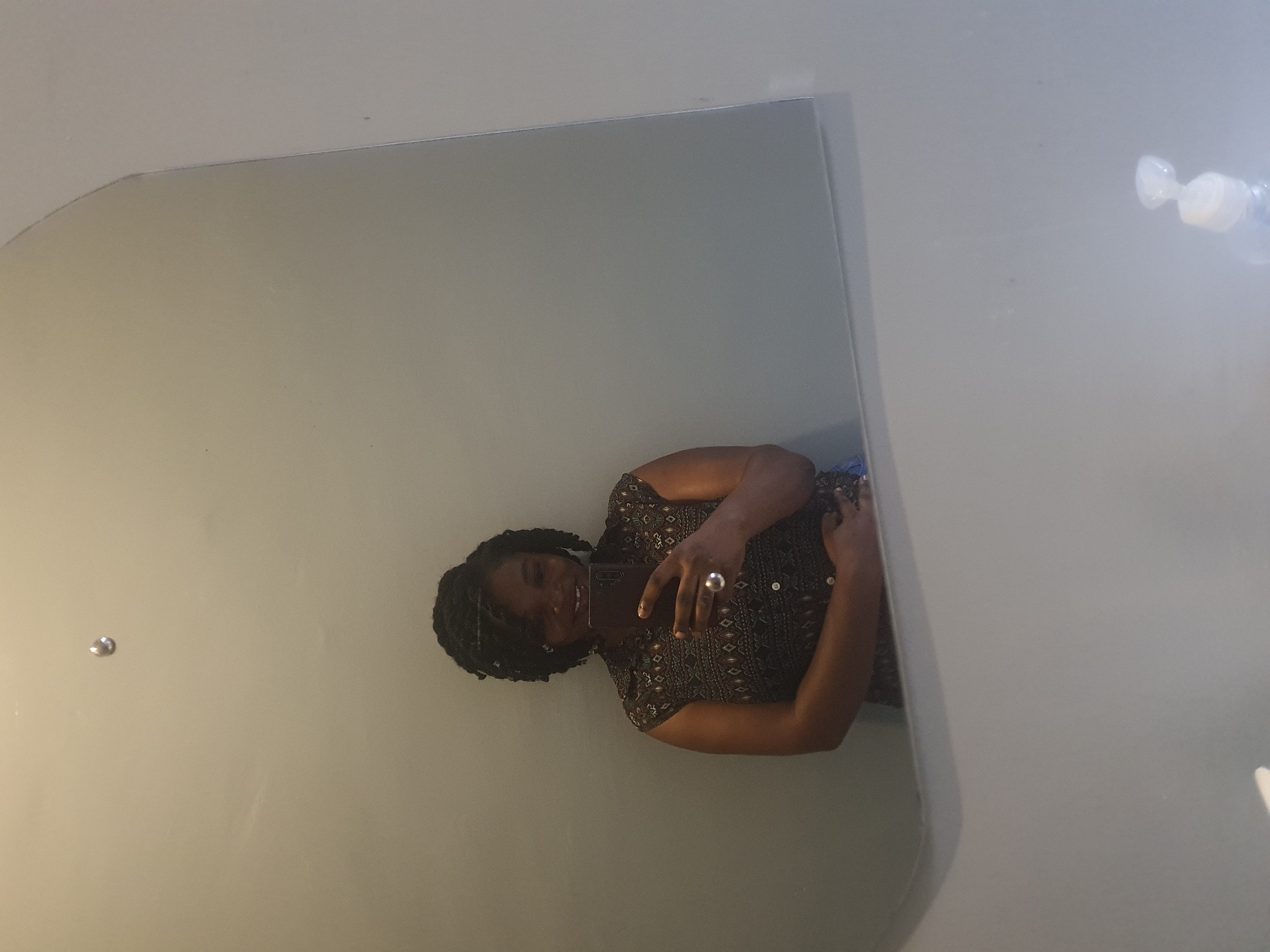 A woman with dark, braided hair takes a selfie in a large, octagonal mirror. She is wearing a dark, patterned sleeveless shirt and a large silver ring on her right index finger. The background is a plain, light gray wall. A bottle with a white pump dispenser is visible at the bottom right of the image. The scene appears to be in a bathroom. The image does not provide any specific visual cues that would indicate its location in Jos, Nigeria.