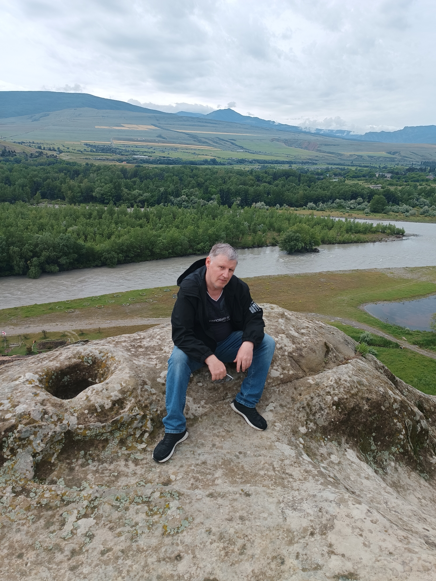 A man is sitting on a rock in Uplistsikhe, Georgia, with a river and mountainous landscape in the background. The man is middle-aged with short grey hair and is wearing a black jacket, blue jeans, and black sneakers. He is holding a pair of sunglasses in his right hand. The rock he is sitting on is large and has a circular indentation. The background shows a wide, grey river flowing through a green valley with a dense forest on one side and fields and mountains in the distance. The sky is overcast. The overall scene is natural and serene.