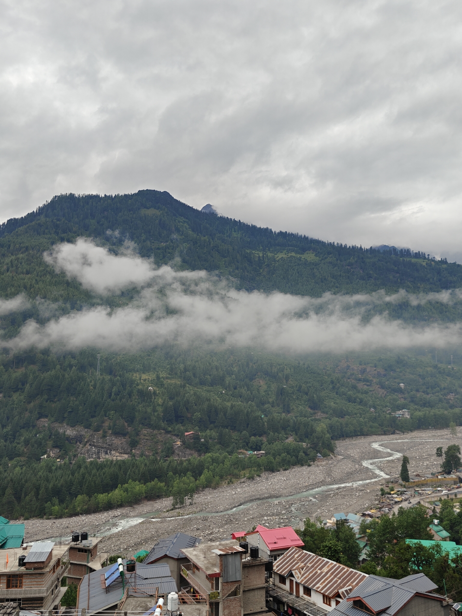 This is a wide-angle, high-angle shot of the town of Vashisht, India, situated in a mountainous region. The sky is overcast with dense, gray clouds. In the mid-ground, a large, tree-covered mountain dominates the scene, with patches of fog or low-lying clouds clinging to its slopes. A river, appearing wide and rocky, flows through the valley below the mountain, with a thin stream of water visible in its center. In the foreground, a dense collection of buildings with pitched roofs, some made of metal and others of tile, are visible, suggesting a residential or commercial area. Several water tanks are mounted on rooftops. The overall impression is of a remote, possibly humid, mountainous environment with a developed settlement in the valley.