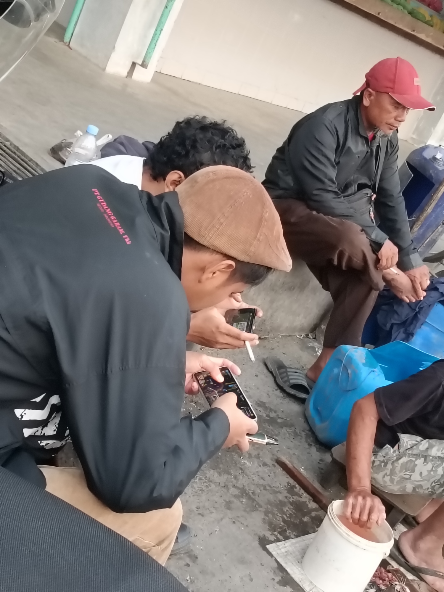 The image depicts a group of men in an outdoor, possibly industrial or service area, with a concrete floor and light-colored walls. In the foreground, a man wearing a black jacket with "PT GUDANG GARAM Tbk" printed on the sleeve in red is looking down at his phone, holding a cigarette in his other hand. To his left, another man in a brown flat cap is also looking at his phone, and appears to be smoking. To the right, a man wearing a red cap and a black jacket is seated and appears to be tending to his foot, possibly bandaging it. In the background, there are large blue containers and a white bucket filled with orange liquid. The overall impression is of a casual gathering of workers during a break. While the location is identified as Kediri City, Indonesia, the specific context within the city is not visually ascertainable.