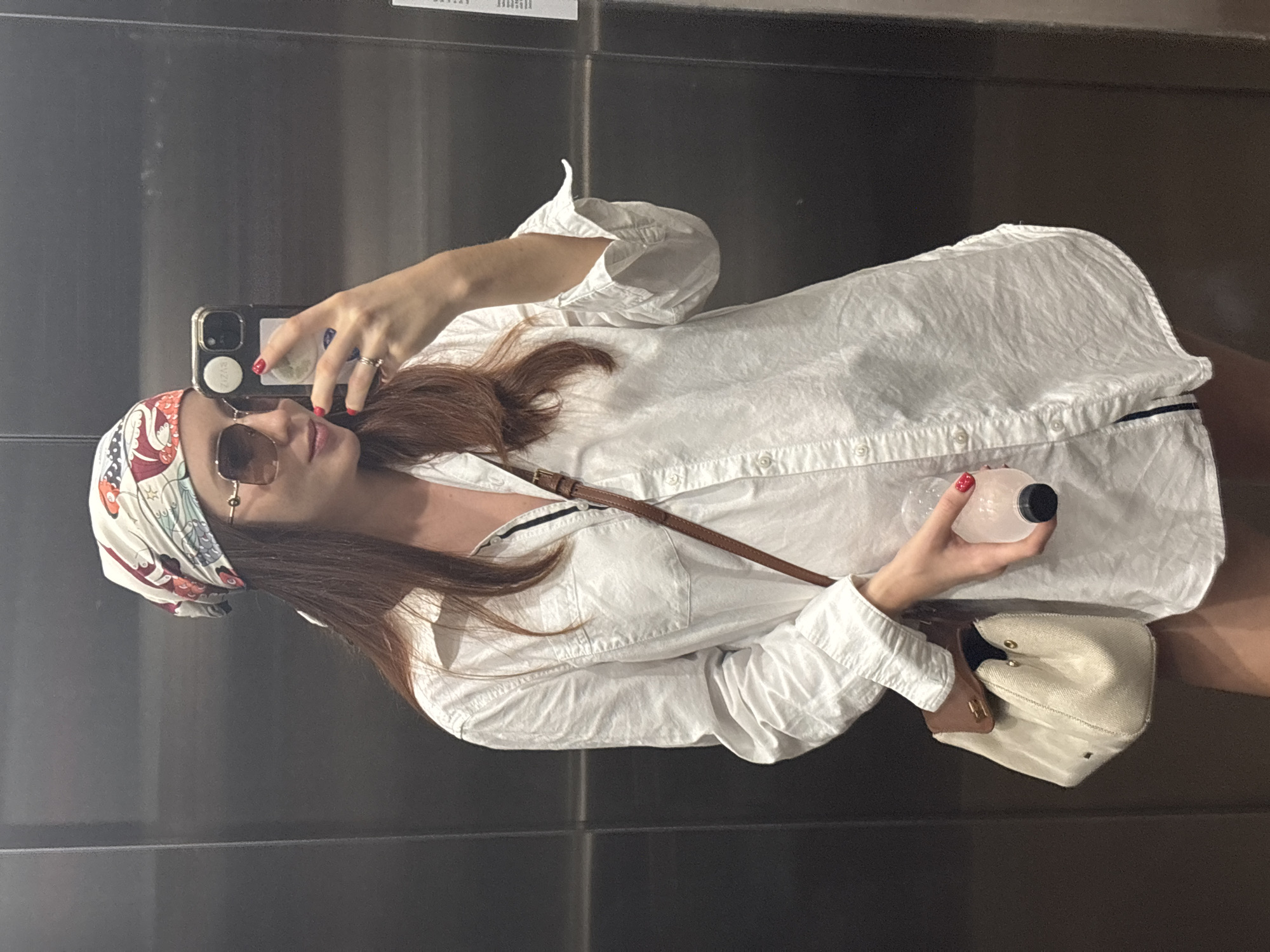 A woman is captured taking a mirror selfie in an indoor setting, likely in Choeng Thale, Thailand. The image is rotated, presenting her horizontally against a dark, reflective background, possibly an elevator or a room with mirrored walls.
The woman has long, straight, reddish-brown hair and is wearing a loose, white button-up shirt, which appears to have a subtle dark trim along a placket or collar edge. She wears a colorful patterned headband or scarf with red, blue, and orange designs on a cream base. Her eyes are concealed behind dark, square-framed sunglasses. Her fingernails are painted red, and a gold-colored ring adorns her left-hand ring finger.
She holds a dark-colored smartphone in her right hand, positioned to capture her reflection. The phone case features white stickers on its back. In her left hand, she holds a small, clear or frosted plastic bottle with a black cap, containing a light-colored liquid. A brown, leather-like strap from a bag crosses her torso, and a light-colored (possibly cream) small handbag with gold-toned hardware is visible near her left side.
The background is composed of dark, uniform panels, suggesting an artificial, indoor environment. The lighting is consistent with indoor conditions, and no specific time of day or weather is evident. The woman maintains a neutral, composed expression typical of someone posing for a photograph. Visible text on the phone case stickers and blurred reflections in the background are not legible.