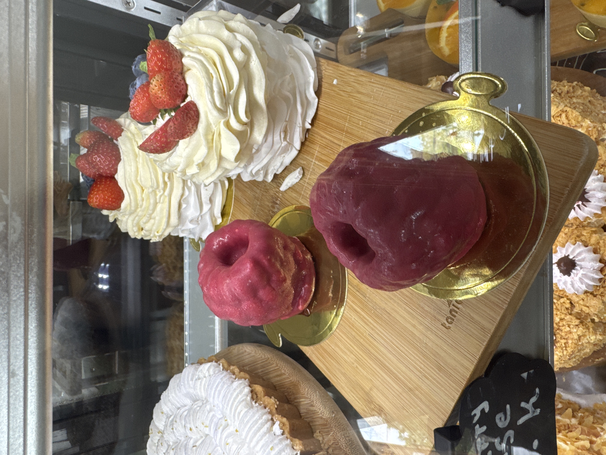 The image shows a close-up view of a display case filled with various desserts. In the foreground, a wooden board holds two bright pink, donut-shaped pastries. To the left, a portion of a white tart with meringue frosting is visible. Behind these, two layered white cakes topped with whipped cream and fresh strawberries and blueberries are arranged. The desserts appear to be for sale in a bakery or patisserie. The overall lighting suggests an indoor setting, likely a commercial establishment. The context of Kuta Selatan, Indonesia, implies this is a local business selling these items.