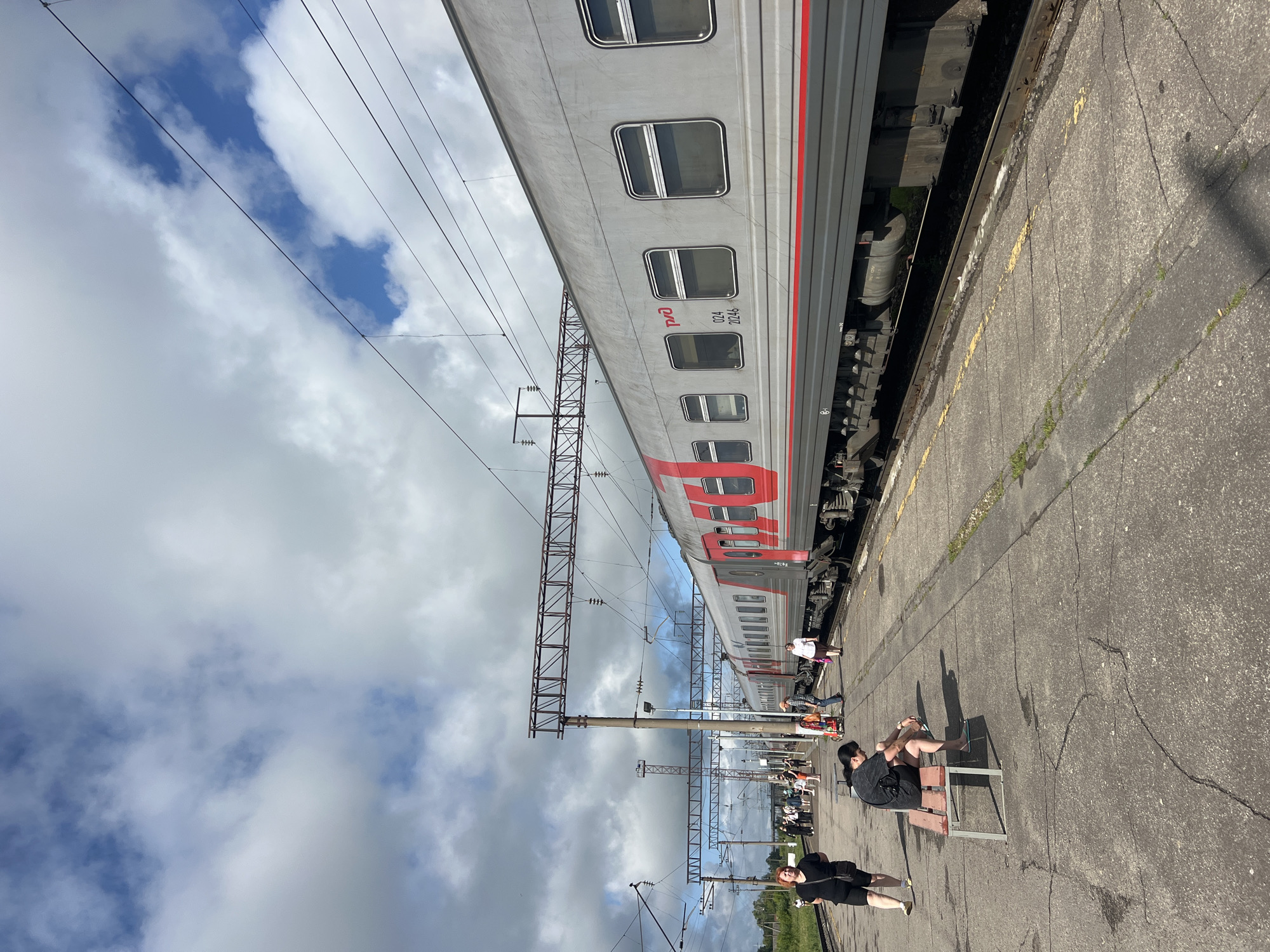 The image depicts a train station platform in Kanash, Russia, under a partly cloudy sky. A modern gray train with red accents is stopped at the platform. Several people are visible on the platform: a woman in a black top and shorts walks away from the camera on the left, a person sits on a bench looking at their phone, and other individuals are scattered throughout the platform, some walking, others standing. The platform is paved with concrete, showing some cracks and stains. Overhead, electrical wires and support structures for the railway are visible. The environment appears to be a functional, open-air train station with greenery visible in the background.