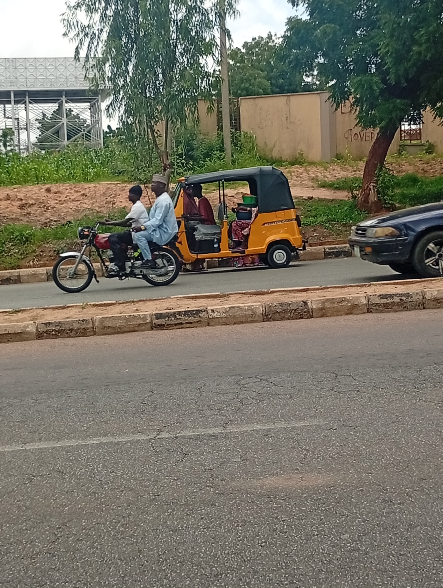 The image shows a street scene in Gombe, Nigeria. A yellow auto-rickshaw is being driven by a man wearing a red shirt, with two passengers behind him. To the left of the auto-rickshaw, a man in a blue traditional outfit and a white cap is riding a red motorcycle, with a younger person behind him. The motorcycle and auto-rickshaw are traveling on a paved road with a raised concrete curb on the right side. In the background, there is a building with the word "GOVER" visible, and dense green foliage with trees. The sky is overcast.