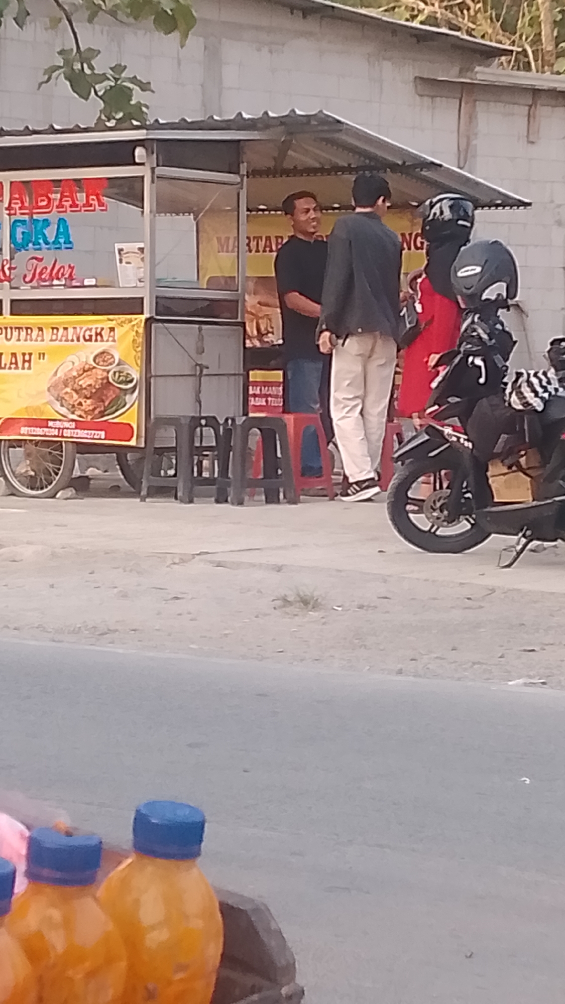 In Karanglangu, Indonesia, a street food stall is visible with colorful signage. Two men are conversing near the stall, one wearing a black t-shirt and the other a grey jacket and beige pants. A woman in a red outfit and a helmet stands behind them. A motorcycle is parked to the right of the people. In the foreground, several orange bottles with blue caps are visible, suggesting drinks are for sale. The stall appears to be selling "Martabak" and "Telur" (egg martabak), with a banner advertising "PUTRA BANGKA ILALAH". The setting is outdoors, likely on a roadside, with a concrete surface in front of the stall.