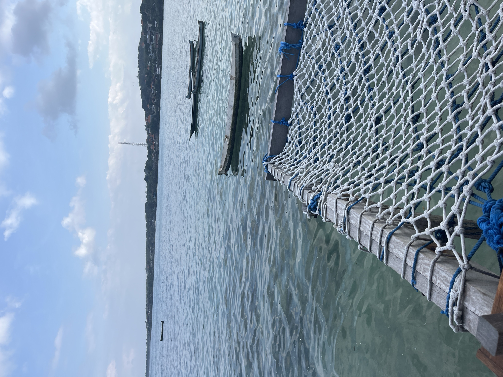 The image shows a close-up of a white net with blue ropes, likely part of a hammock or seating area, extending over calm, clear turquoise water. Several small, traditional wooden boats are floating in the water. In the background, a lush green coastline with scattered buildings and a telecommunications tower can be seen under a partly cloudy sky. The scene appears to be a serene coastal area in Nusa Penida, Indonesia.