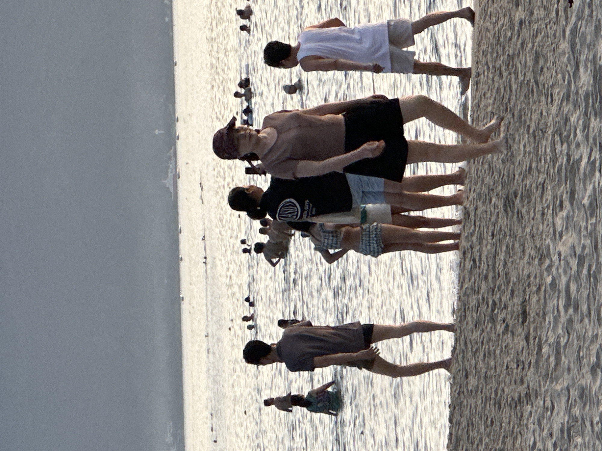 The image depicts a beach scene in Da Nang, Vietnam, with numerous people wading in the water and standing on the shore. In the foreground, several individuals are visible on the sandy beach, with the water lapping at their feet. Further out, groups of people are scattered across the shallow sea, some appearing to be swimming or playing. The background shows a hazy horizon, suggesting a potentially overcast or misty day, with a faint outline of structures or landmass in the distance. The overall atmosphere is one of leisure and recreation, with the natural elements of sand, sea, and sky forming the backdrop for human activity.