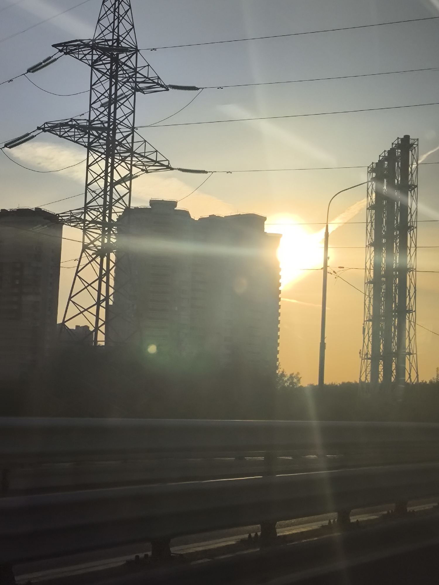 The photograph depicts a cityscape at sunset, featuring a large electrical transmission tower on the left and a tall industrial structure comprised of multiple chimneys on the right. Buildings, likely apartment complexes, stand in the background, their forms partially obscured by the sunlight. A street light is visible to the right of the center, positioned in front of the industrial structure. The foreground is dominated by highway guardrails and a section of the road, suggesting the image was taken from a moving vehicle. A bright glare from the setting sun washes over the scene. The location is labeled as "Unknown, Unknown."