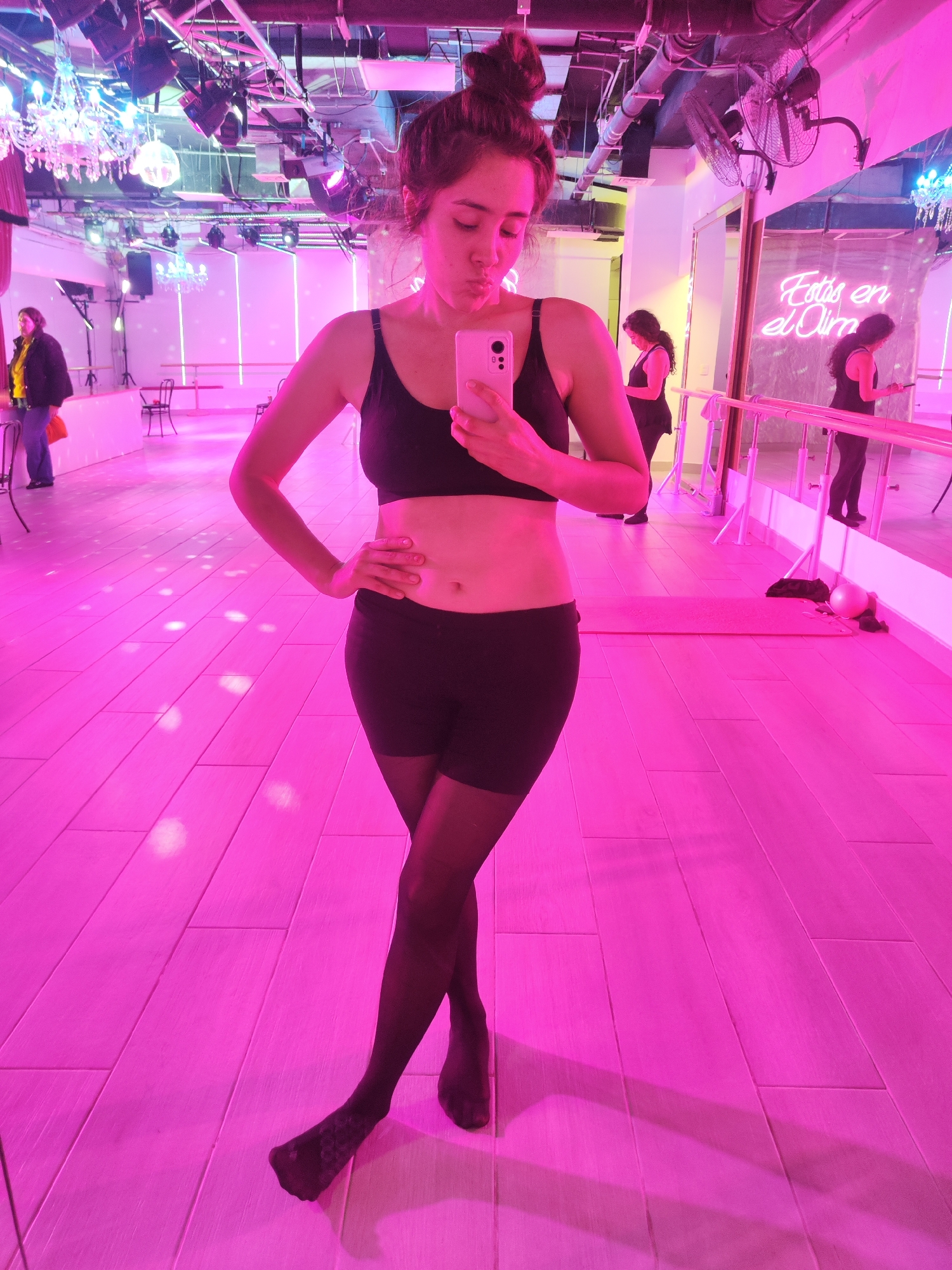 A woman takes a mirror selfie in a dance studio that is bathed in pink light. She is wearing a black sports bra, black shorts, and black sheer pantyhose. Her hair is pulled up in a messy bun. In the background, there are mirrors, ballet barres, and a neon sign that reads "Estás en el Clima". A chandelier hangs from the ceiling, and there are lights and fans. Another woman in a yellow top and black jacket is visible in the background to the left. The floor is made of light-colored wood planks. The overall atmosphere suggests a dance or fitness studio in Santiago, Chile.