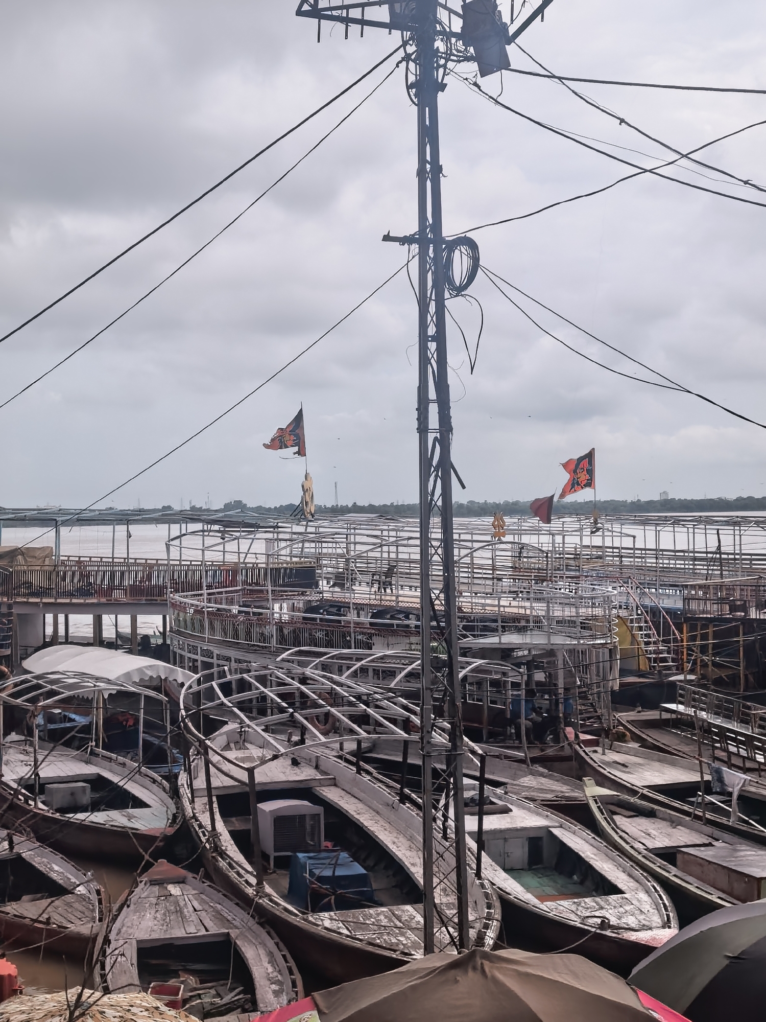 The image depicts a cloudy day scene at the ghats of Varanasi, India. In the foreground, multiple boats are docked, some with white canopies and others with exposed wooden decks and interiors. These boats appear to be of varying sizes and are clustered together. Overhead, a complex network of electrical wires and a tall pole with electrical equipment dominate the upper portion of the frame. In the midground, a more substantial, multi-decked boat or ferry is visible, with its structure made of metal railings and platforms. Two flags are flying from this structure: one is orange with a dark emblem, and another smaller red flag is below it. Beyond this, the Ganges River stretches out, with a bridge or walkway visible in the distance. The background shows a tree line and faint industrial structures against a hazy sky. The overall atmosphere is that of a busy waterfront in a city known for its religious and cultural significance.