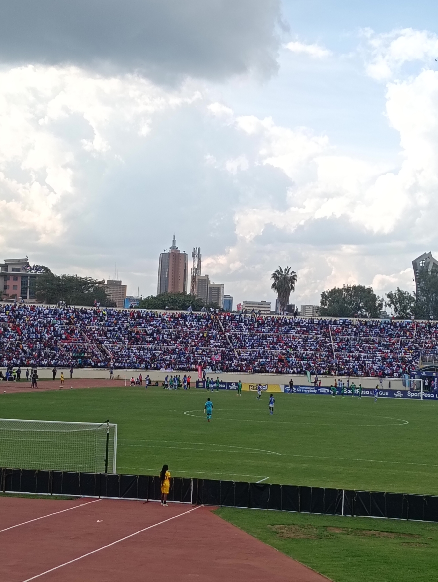 The image shows a soccer game in progress at a stadium in Nairobi, Kenya. The stands are packed with spectators, appearing predominantly dressed in blue and white. Several high-rise buildings are visible in the background skyline. The soccer field is green, and a few players are scattered on the field. A woman in a yellow outfit stands near the edge of the field beside the running track. Signage with the words "SportPesa" is seen along the field's edge. The sky is cloudy.