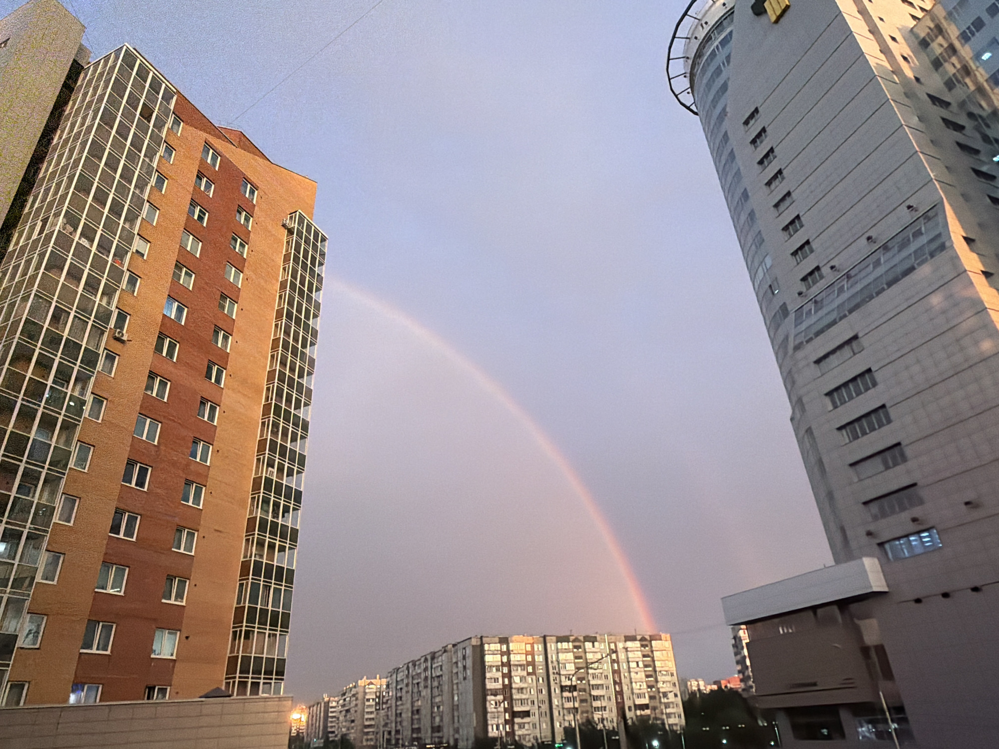 The image captures a cityscape with several apartment buildings and a rainbow in the sky. The dominant subjects are the buildings, which vary in architectural style and height. On the left, a tall, multi-story apartment building with a brick facade and many windows is prominent. To the right, a modern, curved skyscraper with a gray exterior and numerous rectangular windows stands tall. In the background, a long, low-rise apartment complex stretches across the horizon.
A rainbow arcs gracefully across the sky, its colors faintly visible against the pale, overcast sky, suggesting recent or ongoing rain. The lighting indicates it is either dawn or dusk, with a soft, diffused light illuminating the buildings and casting long shadows. The overall mood is serene and perhaps a little melancholic, often associated with the aftermath of a storm and the sight of a rainbow.
There is no visible text in the image. The scene is characteristic of an urban environment, likely a residential area in Krasnoyarsk, Russia, given the provided location context. The focus is on the natural phenomenon of the rainbow set against the backdrop of human-made structures.