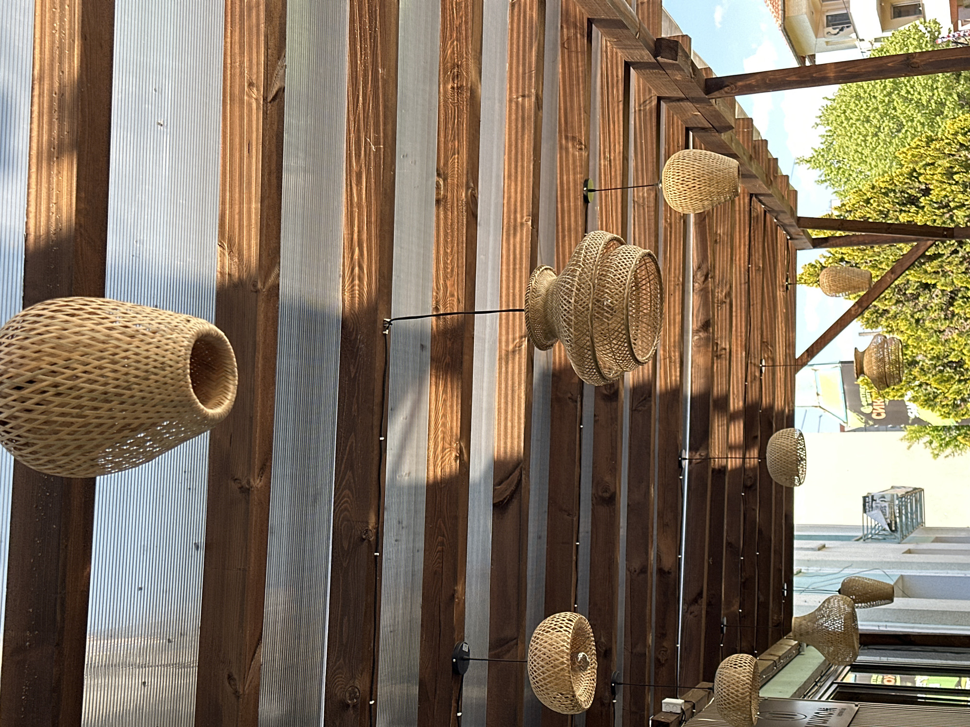 The image shows a series of woven wicker lamps hanging from a wooden structure. The structure appears to be part of a building or outdoor seating area, with alternating wooden and corrugated plastic panels forming the walls. Sunlight casts shadows across the panels, creating a pattern of light and dark. Beyond the structure, there is a glimpse of a city street with buildings and trees, suggesting an urban location. The objects appear to be stationary. There are no visible people.