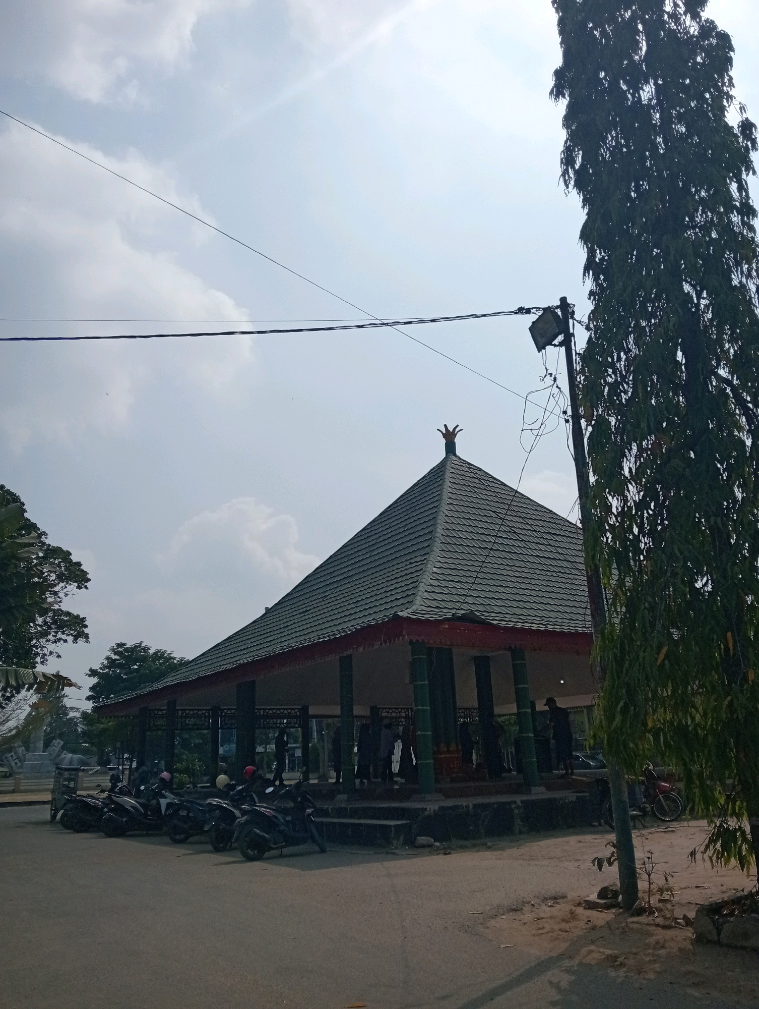 In Pringsewu, Indonesia, an outdoor scene features a traditional open-sided pavilion with a dark green, multi-tiered roof supported by pillars. Several people are visible under the pavilion, some appearing to be interacting. A row of parked motorcycles, predominantly black, is situated on the left side of the image, facing the pavilion. A tall, leafy tree dominates the right side of the frame, its foliage casting shadows. A utility pole with a spotlight and complex wiring stands near the tree and pavilion. The ground is a mix of asphalt and dirt. The sky above is bright and partly cloudy, with visible power lines crisscrossing the frame. The overall impression is a public gathering or recreational space.