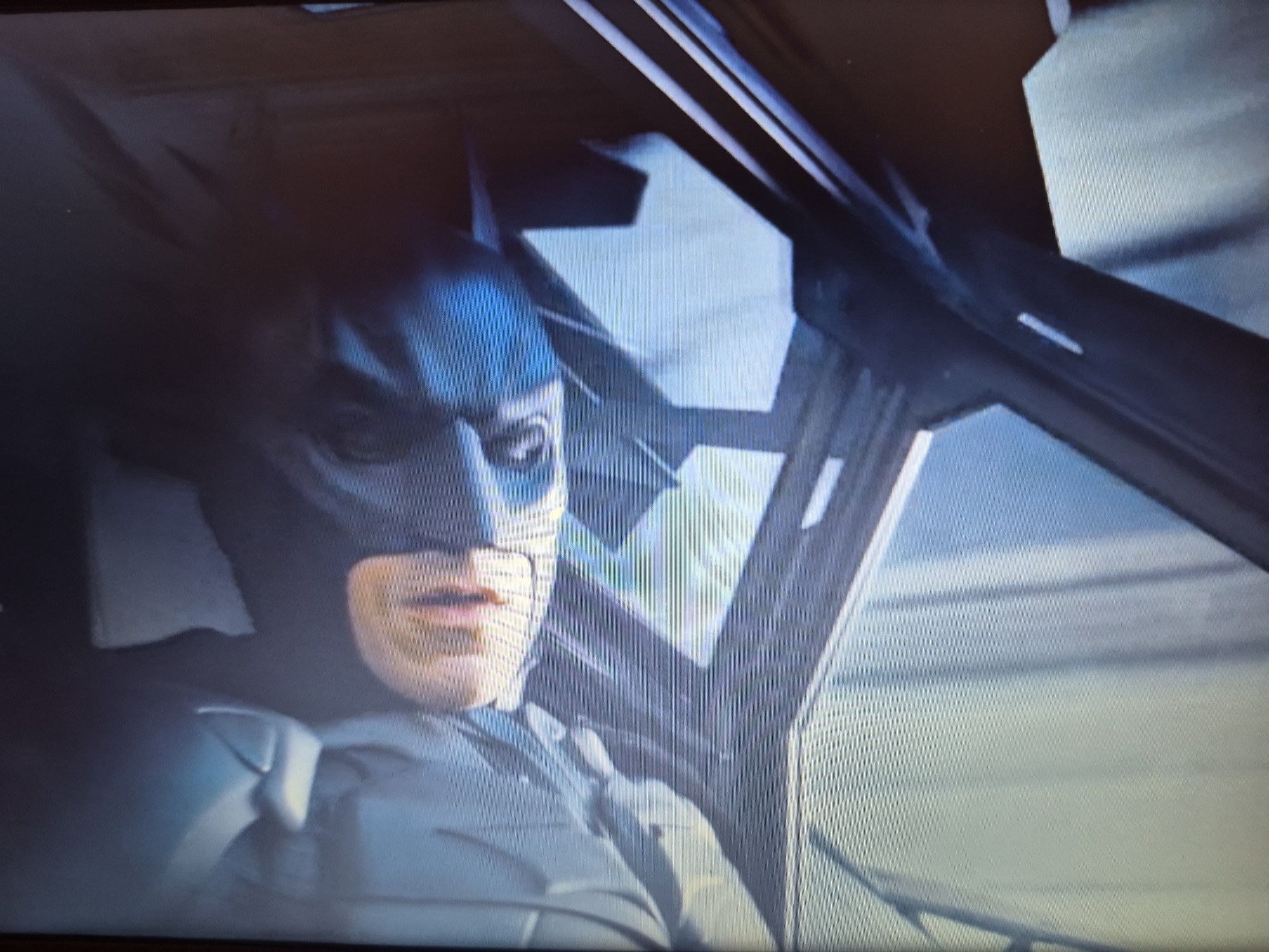 The image shows a close-up of Batman, wearing his mask and cowl, looking out of a vehicle. He is wearing his armored suit, which is predominantly dark gray with some lighter gray or blue accents on the shoulders. The background appears to be the interior of a vehicle, with a window or windshield showing a blurry, light-colored environment, possibly a cityscape or sky. The perspective suggests he is the driver or a passenger in the front of the vehicle. There is no discernible information within the image that would indicate a location in Sandwell, United Kingdom.