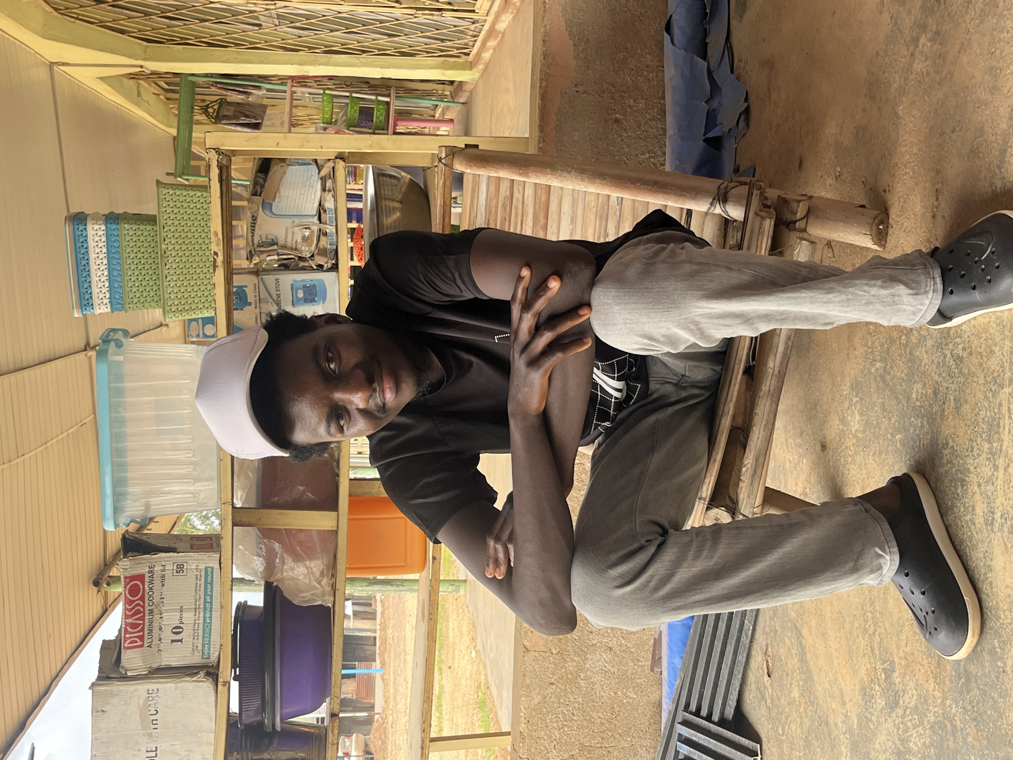 A young Black male wearing a black t-shirt, grey pants, a white baseball cap, and black clogs sits on a bamboo chair. He has his arms crossed and is looking towards the camera. The background shows shelves filled with various items, including containers, boxes, and baskets. The scene appears to be outdoors, possibly a market or storage area, with a dirt floor and a corrugated roof overhead.