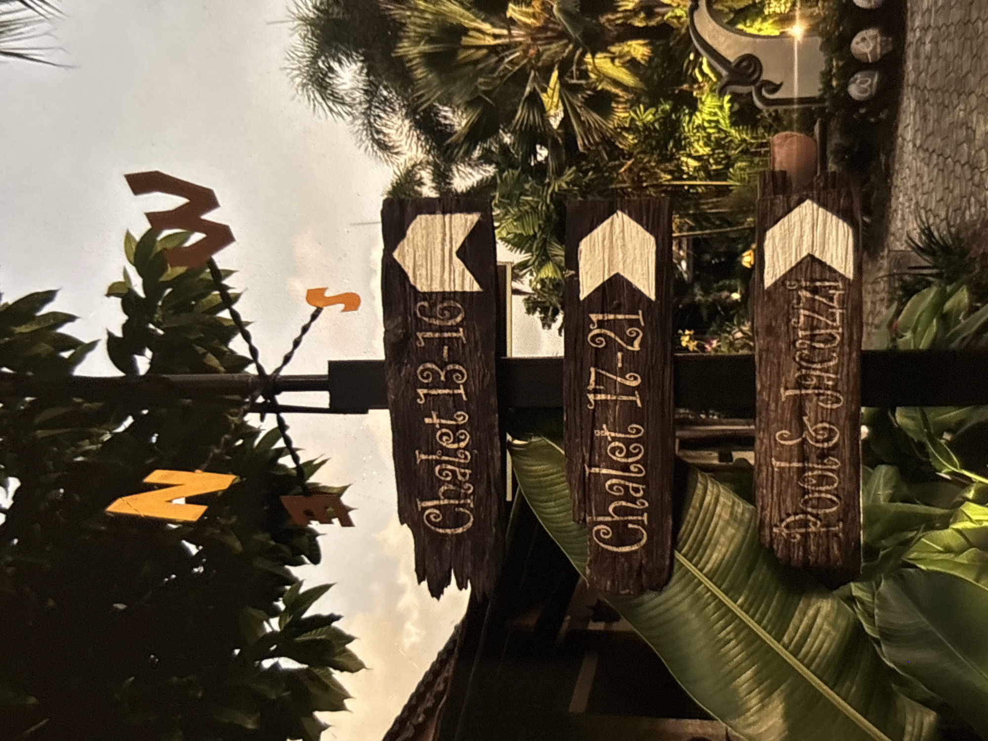 A close-up, vertical shot shows a dark wooden signpost with three signs pointing to the right. The top sign reads "Chalet 13-16", the middle sign reads "Chalet 17-21", and the bottom sign reads "Pool & Jacuzzi". All text is in a decorative, light-colored font. The signs are attached to a central black pole. Above the signs, a directional arrow, possibly part of a weather vane, is visible against a bright, overcast sky. Orange letters "N" and "W" are also visible in the foliage above. Lush green tropical foliage, including large banana-like leaves and palm fronds, frame the signpost on the left and right. In the background, palm trees and a softly lit archway suggest an outdoor resort or garden setting, consistent with the resort atmosphere of Chalong, Thailand. The ground in the foreground is paved with interlocking stones. The overall impression is that of an entrance or directional marker within a tropical resort.