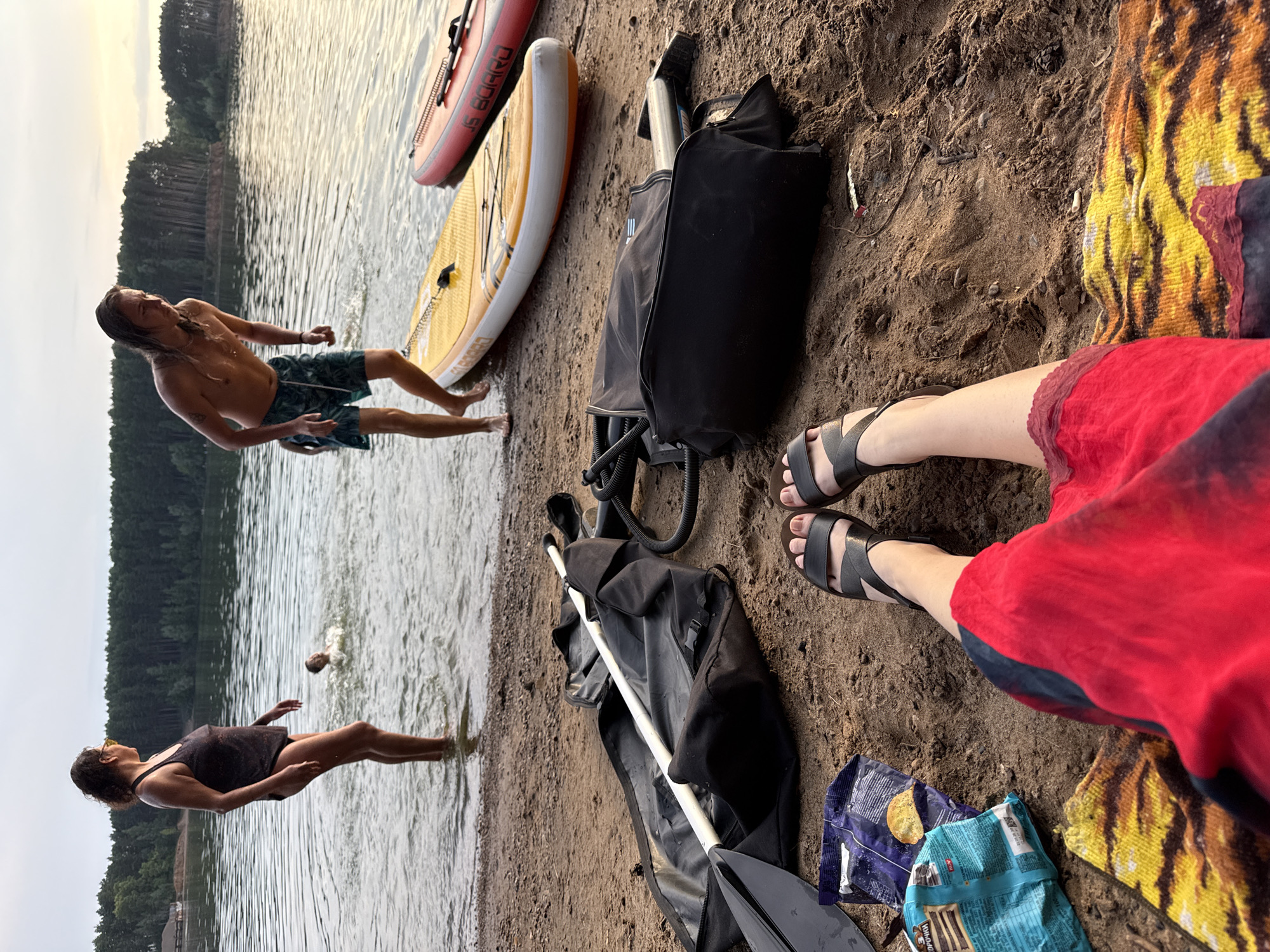 In the city of Unknown, Unknown, on a sandy beach beside a body of water, a group of people are enjoying a leisurely day. In the water, a woman in a black swimsuit stands waist-deep, looking out at the scene. A child is splashing nearby. A young man with long hair, wearing patterned swim shorts, is standing by the water's edge, preparing to enter the water, with a paddleboard beside him. Another paddleboard, red in color, lies on the sand next to it. In the foreground, a pair of black sandals are worn by a person lying on a towel with a vibrant tiger print. A red and black garment is also visible. Next to the towel are bags, a paddle, and two packets of snacks. The background features a dense line of trees and a calm body of water, likely a lake. The sky is overcast, suggesting either early morning or late afternoon.