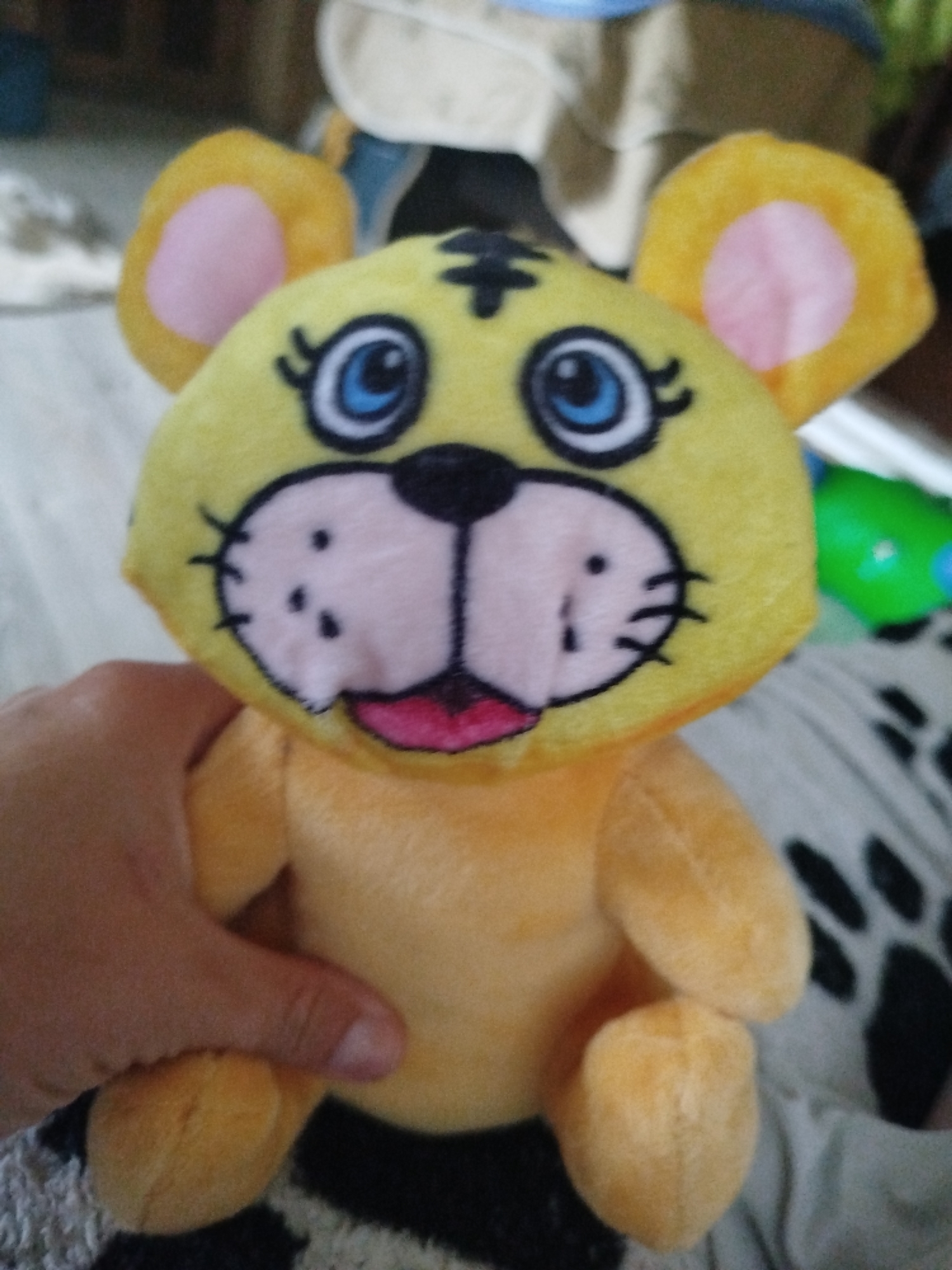 A close-up, slightly blurred, vertically oriented photograph captures a stuffed toy tiger held by a person's hand. The toy is predominantly yellow with pink inner ears and a light pink muzzle. It has large blue eyes with black eyelashes, black stripes on its forehead, a black nose, and a pink mouth. The toy's body is a lighter shade of orange-yellow. The hand appears to be gripping the toy around its lower body, with fingers visible on the left side of the frame. The background is out of focus, showing a patterned surface that appears to be fabric, possibly a blanket, with black and white paw prints. To the right, a bright green object with blue details is partially visible. The scene is indoors, and based on the provided location information, it is in Krasnousolskij, Russia. The overall lighting suggests an indoor environment, possibly during daytime, though the lighting conditions are not clearly discernible due to the focus and blur.