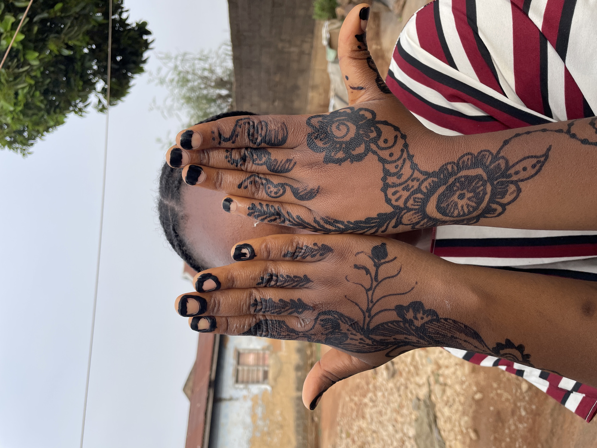 A medium shot shows a person with brown skin and black nails covering their face with their hands, which are adorned with intricate black henna designs. The person wears a striped shirt with burgundy, white, and black stripes. The background includes a building with a brown roof and a window with closed shutters. A brick wall and greenery are also visible. The image appears to be taken outdoors in the city of Jos, Nigeria.