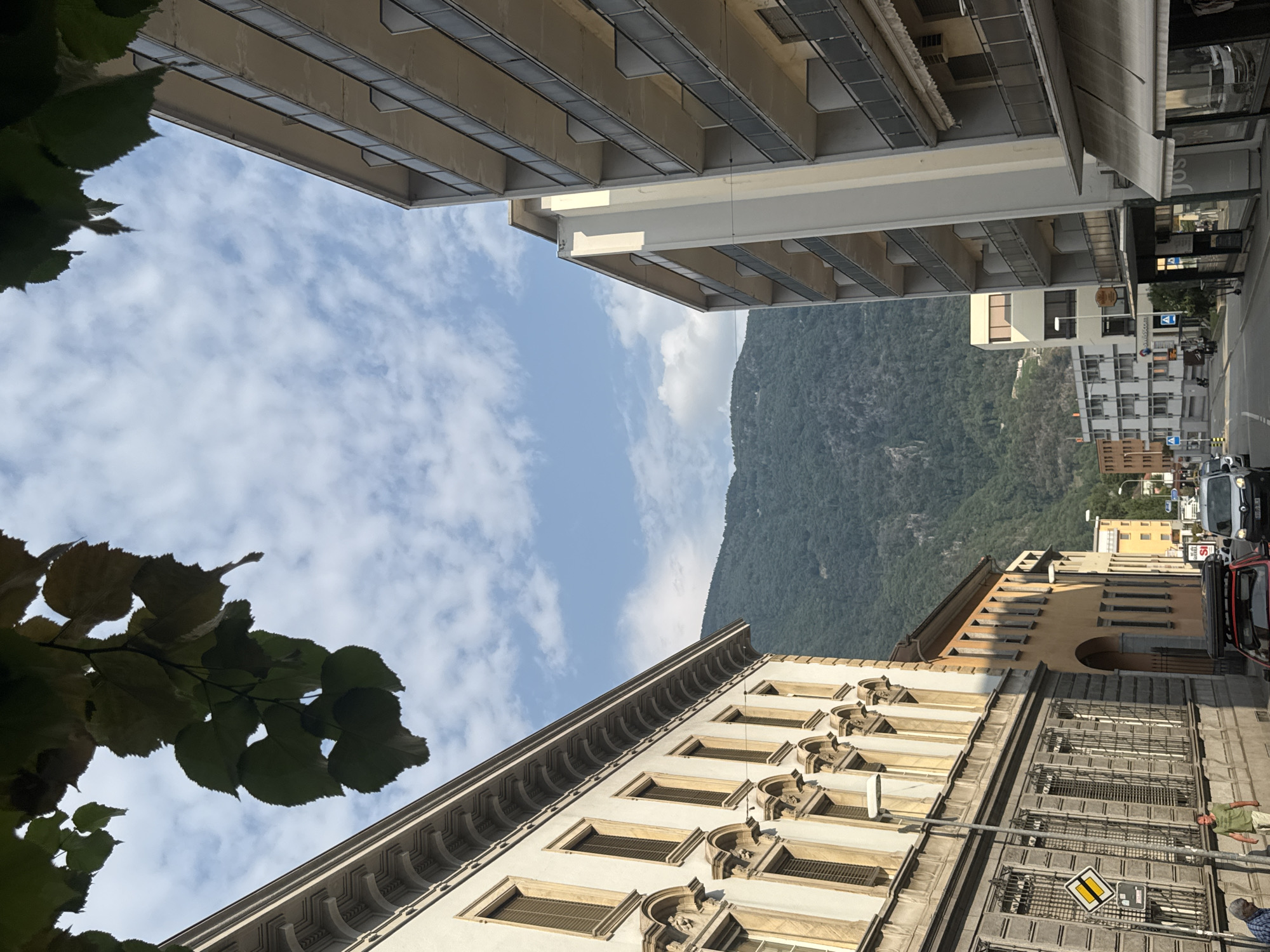 This is an eye-level, medium-shot, vertical-format photograph taken on a sunny day in Bellinzona, Switzerland. The view is looking up a street lined with multi-story buildings on either side, with a lush, green mountain in the background. The sky is a clear blue with scattered white clouds.
On the left side of the street, a light-colored building with classical architectural details, including decorative molding and arched windows, dominates the foreground. The windows have brown shutters. Below this building, a grey stone building with barred windows and a sidewalk in front of it is visible. A street lamp is positioned on the sidewalk, and two men are standing on the sidewalk. A yellow and black traffic sign is mounted on a pole.
On the right side of the street, a modern, multi-story apartment building with balconies is visible. The building is lighter in color, with glass railings on the balconies. Further down the street, other buildings of varying heights and architectural styles are present.
The street itself is paved and appears to be a typical urban street with parked and moving vehicles. The scene suggests a bustling city environment with a strong natural backdrop.