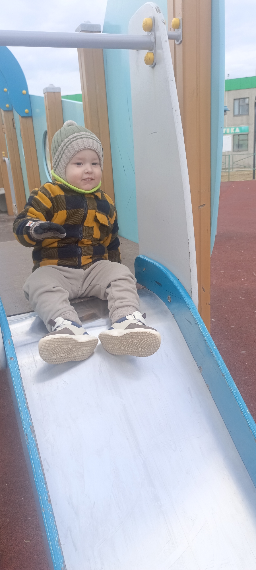 A young child is seated at the top of a metallic slide with blue side railings on an outdoor playground. The child, appearing to be a toddler or preschooler, looks towards the camera with a slight smile, suggesting contentment.
They are dressed for cool weather, wearing a light green and beige striped beanie hat with a pom-pom, a bright green neck warmer, a yellow and black plaid zip-up jacket, light brown pants, and light-colored sneakers with chunky soles. They also have gloves or mittens on their hands.
The playground structure behind and around the child consists of light blue panels, wooden posts, and white support beams with yellow cap fasteners. In the background, beyond the playground, a light green building with windows is visible, and another building features text that reads "АПТЕКА" (Pharmacy) in Russian. The ground of the playground appears to be a reddish-brown safety surface. The lighting suggests an overcast daytime sky. The location is identified as Markovskiy, Russia.
