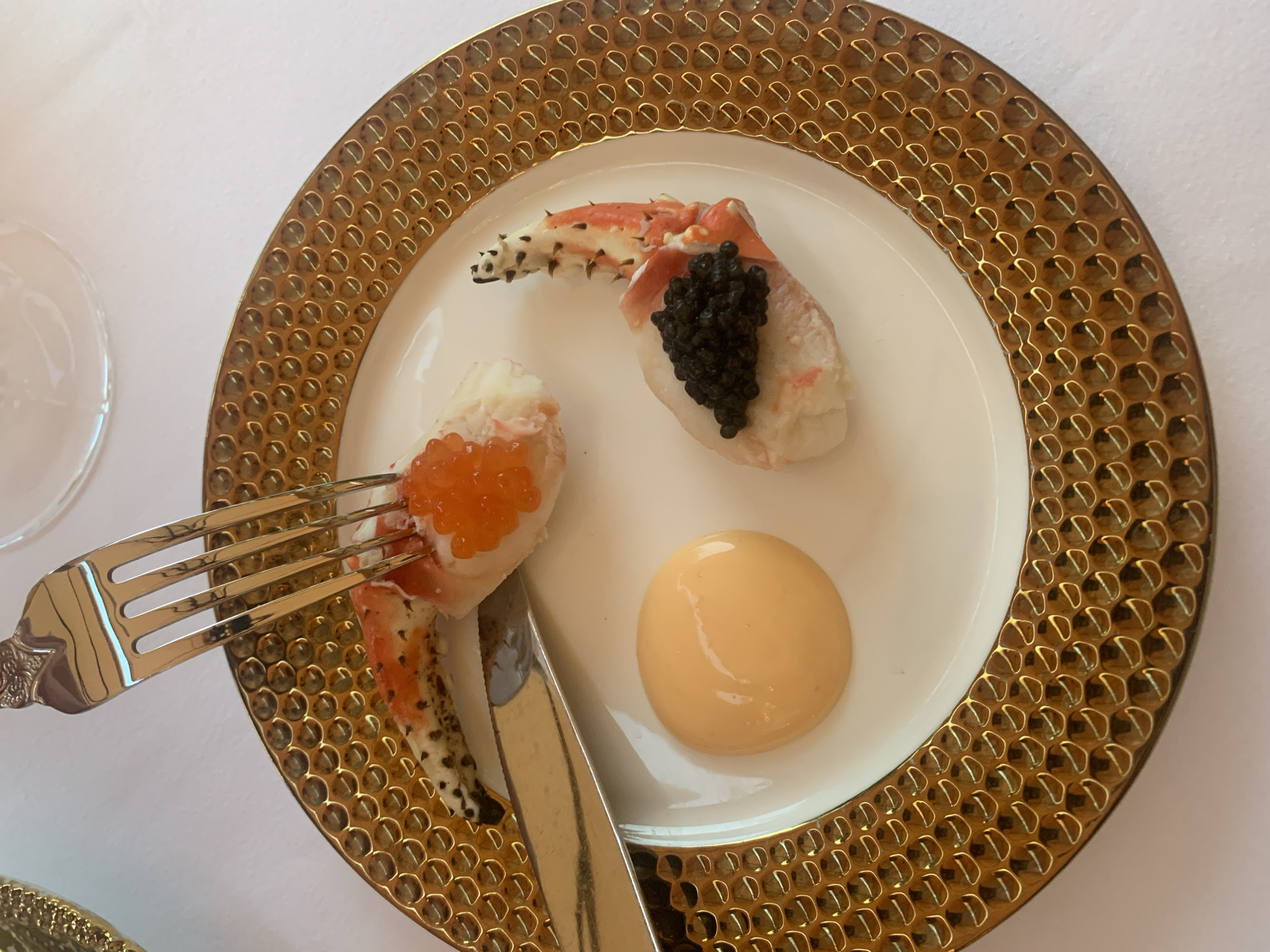 An elegant gold-rimmed white plate holds a delicate seafood appetizer. Two pieces of king crab leg, one adorned with bright orange tobiko (flying fish roe) and the other with glossy black caviar, are arranged on the plate. A dollop of creamy orange sauce sits to the left of the crab. A silver fork is poised above one of the crab legs, suggesting it is ready to be eaten, and a silver knife rests on the plate. The table is covered with a white tablecloth. The scene appears to be in a high-end restaurant setting. No specific details about the location in Moscow, Russia are visible in the image.