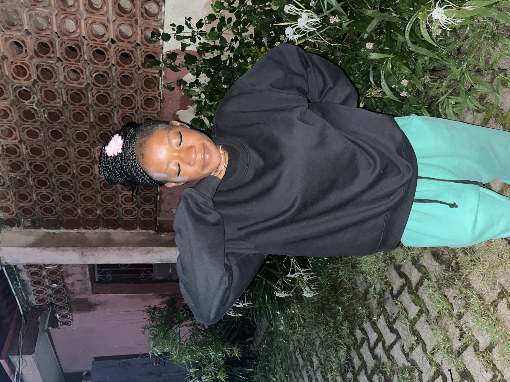 A young Black woman with braided hair, adorned with a pink flower, stands with her eyes closed and head tilted back slightly. She is wearing a black sweatshirt and bright green sweatpants. Her left arm is raised, with her hand resting on the back of her neck, and her right arm is bent at the elbow with her hand on her hip. The background features a building with decorative patterned brickwork and a pink-toned wall. Lush green foliage, including some white flowers, surrounds the woman, and the ground is paved with interlocking bricks interspersed with grass. This scene appears to be an outdoor setting at dusk or night, possibly in a residential area within Jos, Nigeria, given the architectural style and vegetation.