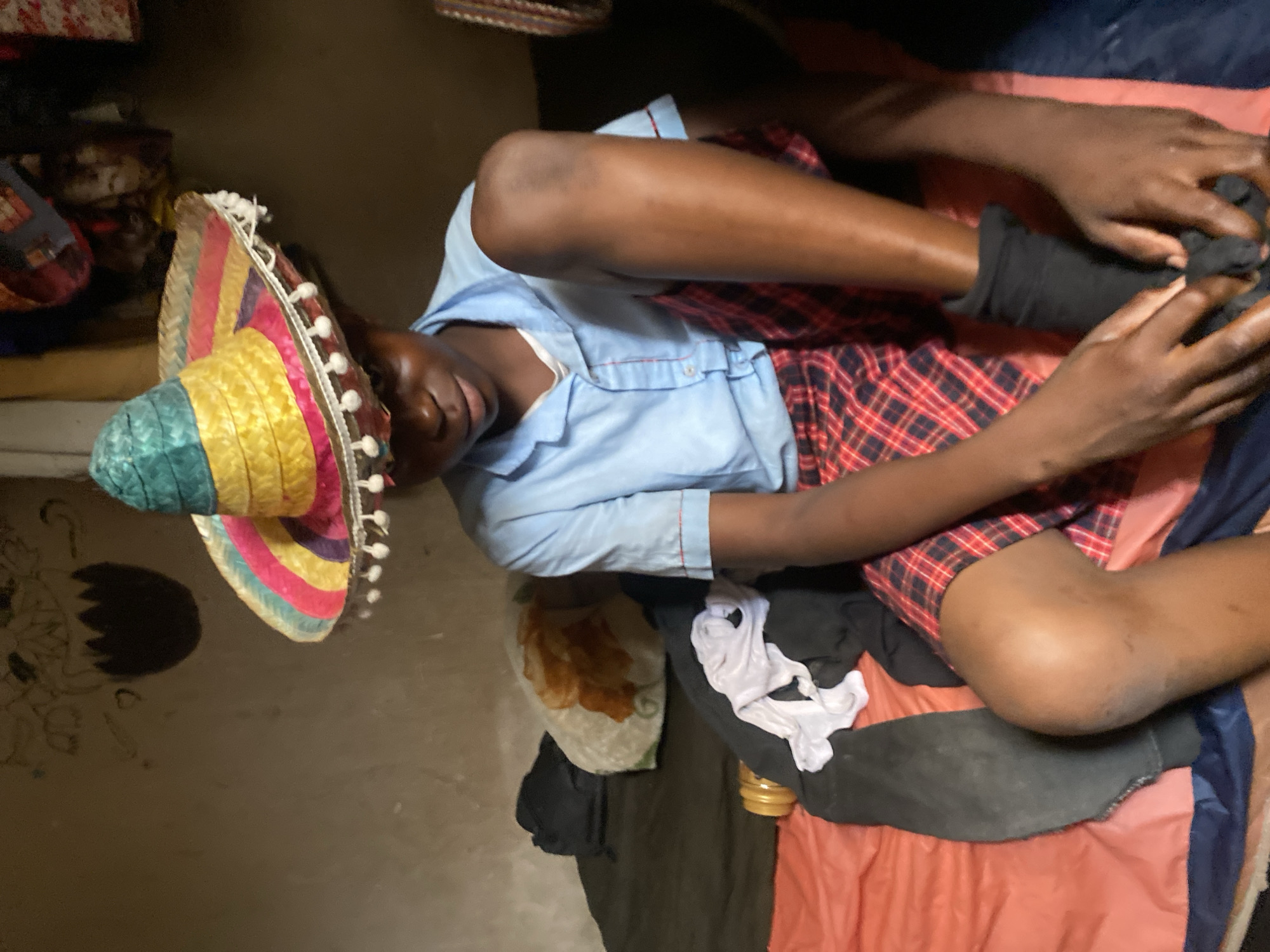 A person wearing a sombrero, a light blue shirt, and a red plaid skirt is lying on a red and blue surface. The person is touching their left foot, which is covered in a black sock. There is clothing scattered around them, including black and white fabric. The background appears to be an interior space with items stacked in the corner. The city of Bukuru, Nigeria, is the assumed location.