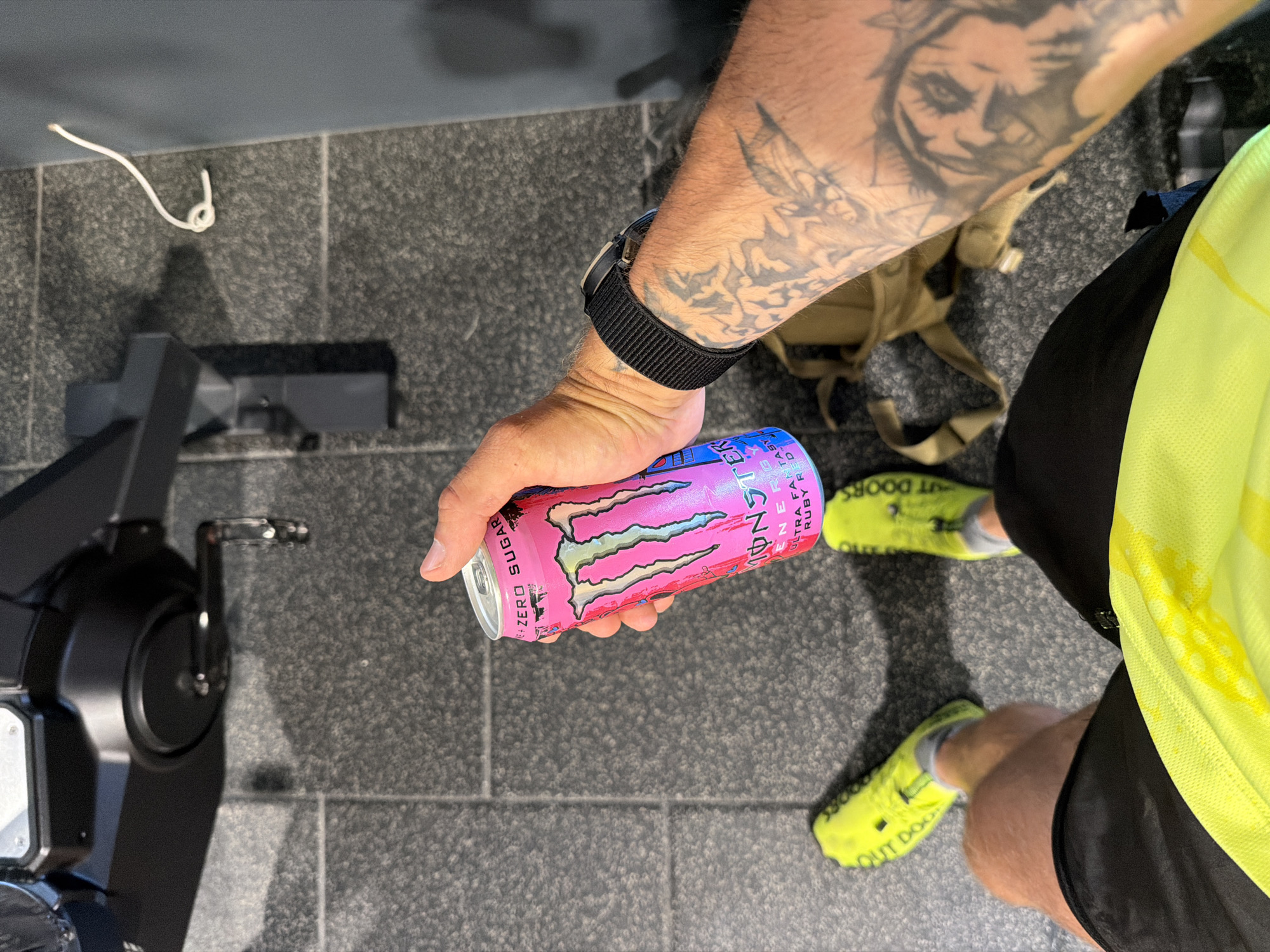 The image shows a person's hand holding a pink and blue can of Monster Energy drink, with the label visible. The person is wearing a black watch on their left wrist, and has tattoos on their left arm. The person is also wearing bright yellow athletic shoes with black writing on them, black shorts, and a neon yellow shirt. The background appears to be a gym, with a piece of exercise equipment and a textured floor. There is a tan backpack on the floor. The image is taken from a first-person perspective. The location is not explicitly stated in the image, but if the context were Plovdiv, Bulgaria, it would be described as a gym within that city.