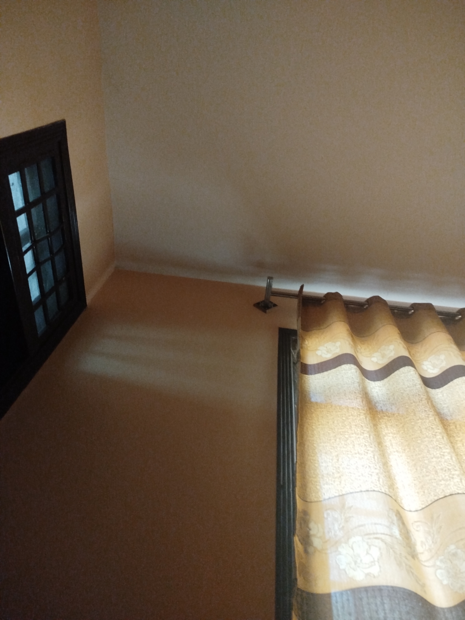 The image shows an interior view of a room. In the foreground on the left is a dark-framed window with multiple panes. To the right, a patterned curtain, primarily in shades of yellow and brown, hangs from a curtain rod. The walls are painted a light, neutral color. The lighting suggests it may be daytime, with some shadows visible on the floor. The scene is devoid of any people or visible activity. The location is specified as Lassan, Pakistan.