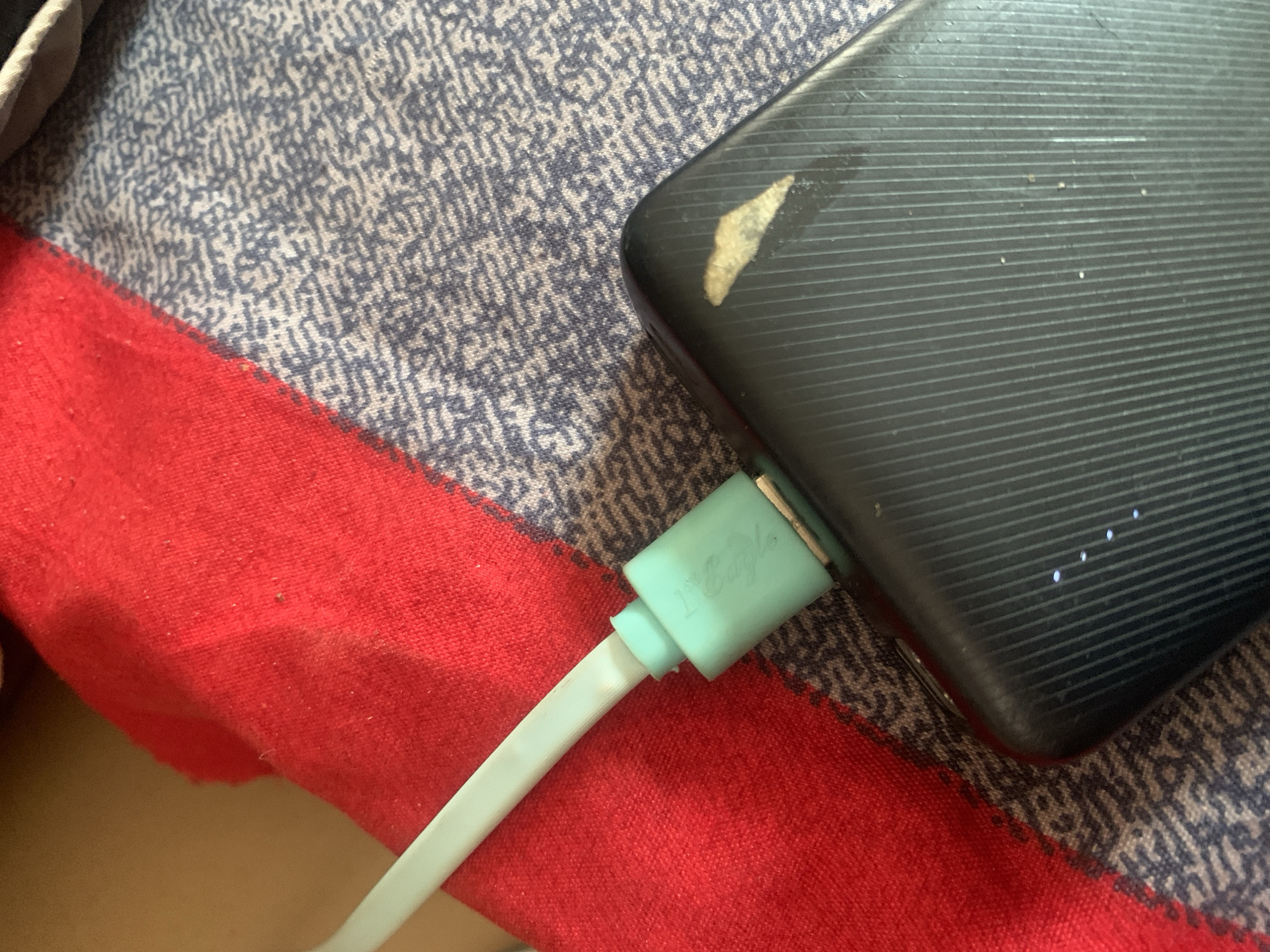 A close-up, high-angle shot shows a light blue charging cable plugged into a black power bank. The power bank has a ribbed texture and a light-colored stain on its surface. The cable is connected to a teal-colored adapter, which is partially visible. The objects are resting on a surface covered by a red cloth on the left and a patterned blue and white fabric on the right. The scene is devoid of any discernible city or geographical indicators, making it impossible to determine its location in Jauro Yinu, Nigeria, or any other place.