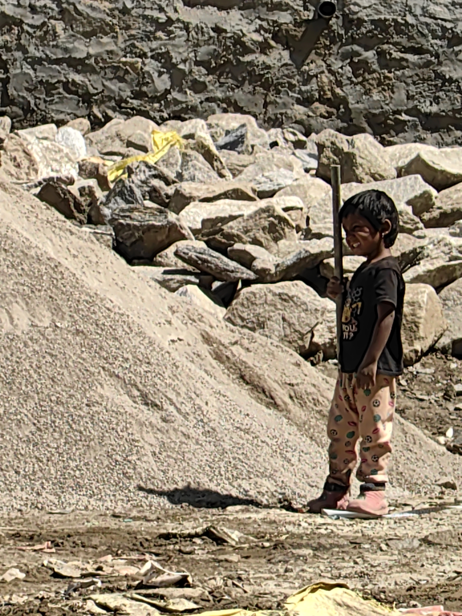 A child stands outdoors in Vashisht, India, near a construction site. The child, wearing a black t-shirt and patterned pants, holds a wooden stick. The background consists of a stone wall and piles of gravel and rocks. The ground is covered in dirt, debris, and some yellow fabric.