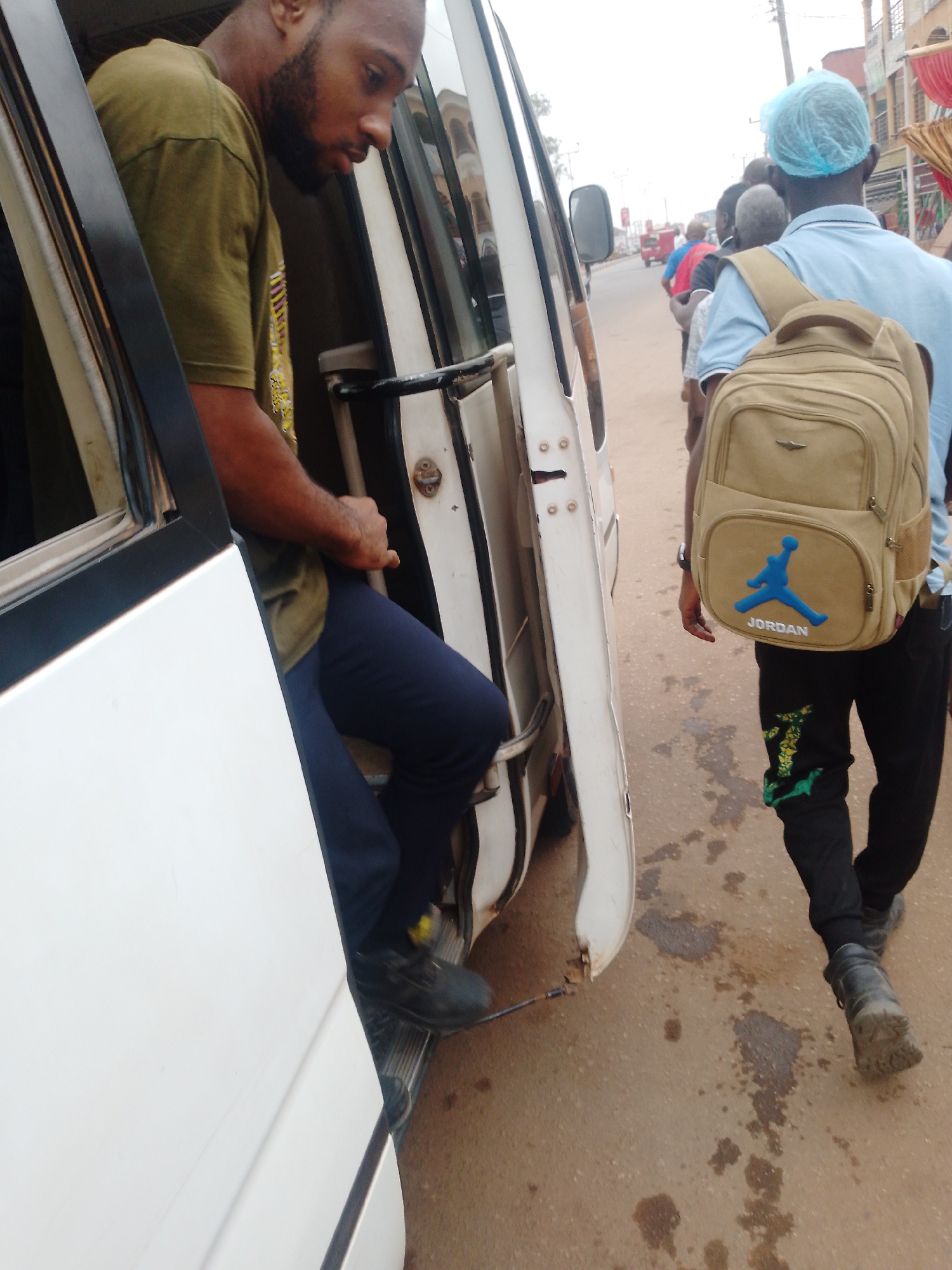 A man with a beard wearing a green shirt and blue pants is getting out of the passenger door of a white van. The van is on a street in Sabon Tasha, Nigeria. There is a line of people outside the van. One man in the line is wearing a blue shirt and has a tan backpack with a blue Jordan logo. The road is paved and shows signs of dirt and minor stains. A building is visible in the background.
