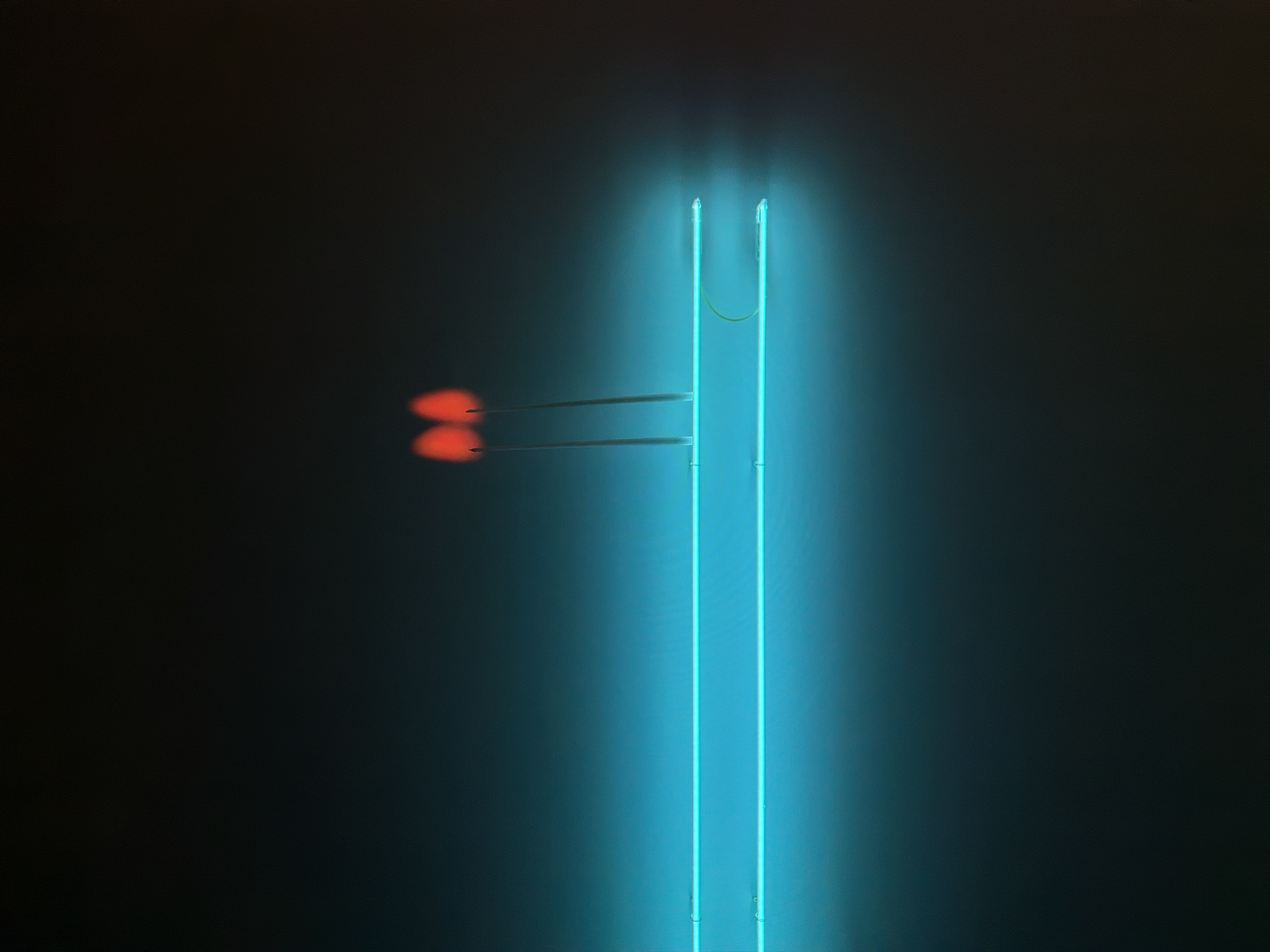 An indoor view shows what appears to be the neon installation "Wall Arrow" by Bruce Nauman on display at the MOCA (Museum of Contemporary Art) in Bangkok, Thailand. Two red neon tubes on the left end like arrowheads pointing towards a taller vertical U-shaped blue neon tube.