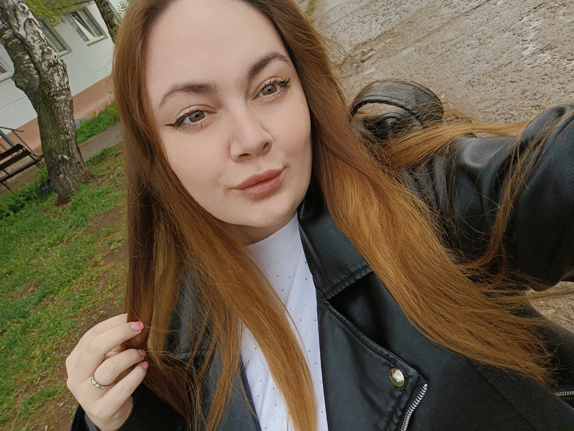 A young woman is taking a selfie. She has light skin, brown eyes with winged eyeliner, and long auburn hair. She is wearing a black leather jacket over a white shirt. She has a ring on her left hand. In the background, there is a birch tree, a building with windows, and a patch of green grass. The location is likely in Chaikovsky, Russia, based on the prompt. The ground appears to be pavement or concrete.