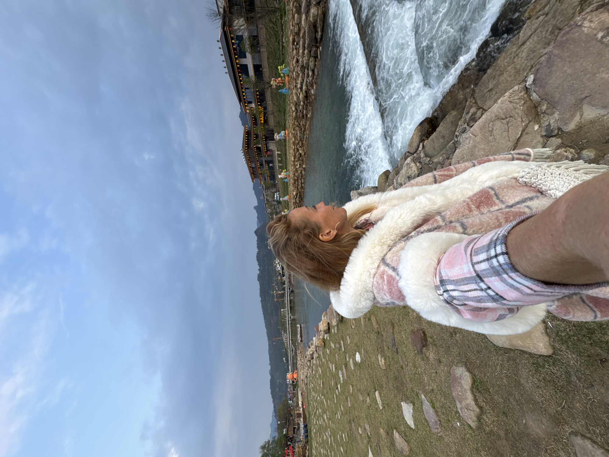 The image captures a forensic wide shot of a woman taking a selfie on a rocky riverbank in the city of Wulingyuan, China, at dusk.
**People:** A woman is positioned in the foreground, her right arm extended towards the camera, holding it to capture the scene. She has light-colored hair, parted on the side, and wears small gold hoop earrings. She is dressed in a thick, pink and white plaid jacket with a prominent white faux fur collar and lapels. Her head is turned to her right, looking out over the river and the opposite bank.
**Environment and Subjects:**
The woman stands on a rugged embankment composed of large, irregular stones, which lines the edge of a river. To her immediate right, the river flows over a visible weir or small cascade, creating turbulent, white-capped water. Beyond the river, on the opposite bank, several multi-story buildings of traditional Chinese architectural style are visible, featuring dark tiled roofs and warm-toned lights illuminating their eaves and windows. In front of these buildings, lining the river, are several distinct, colorful, illuminated animal-shaped sculptures, including what appear to be mice and a large orange octopus further to the left.
To the left of the woman, the embankment transitions into a grassy area interspersed with flat, irregular stones. In the distant background, a bridge spans the river, and multiple construction cranes are visible on the far bank. Undulating mountains rise beyond the urban development, silhouetted against an overcast sky displaying shades of light blue and grey, indicative of either early morning or evening light.
