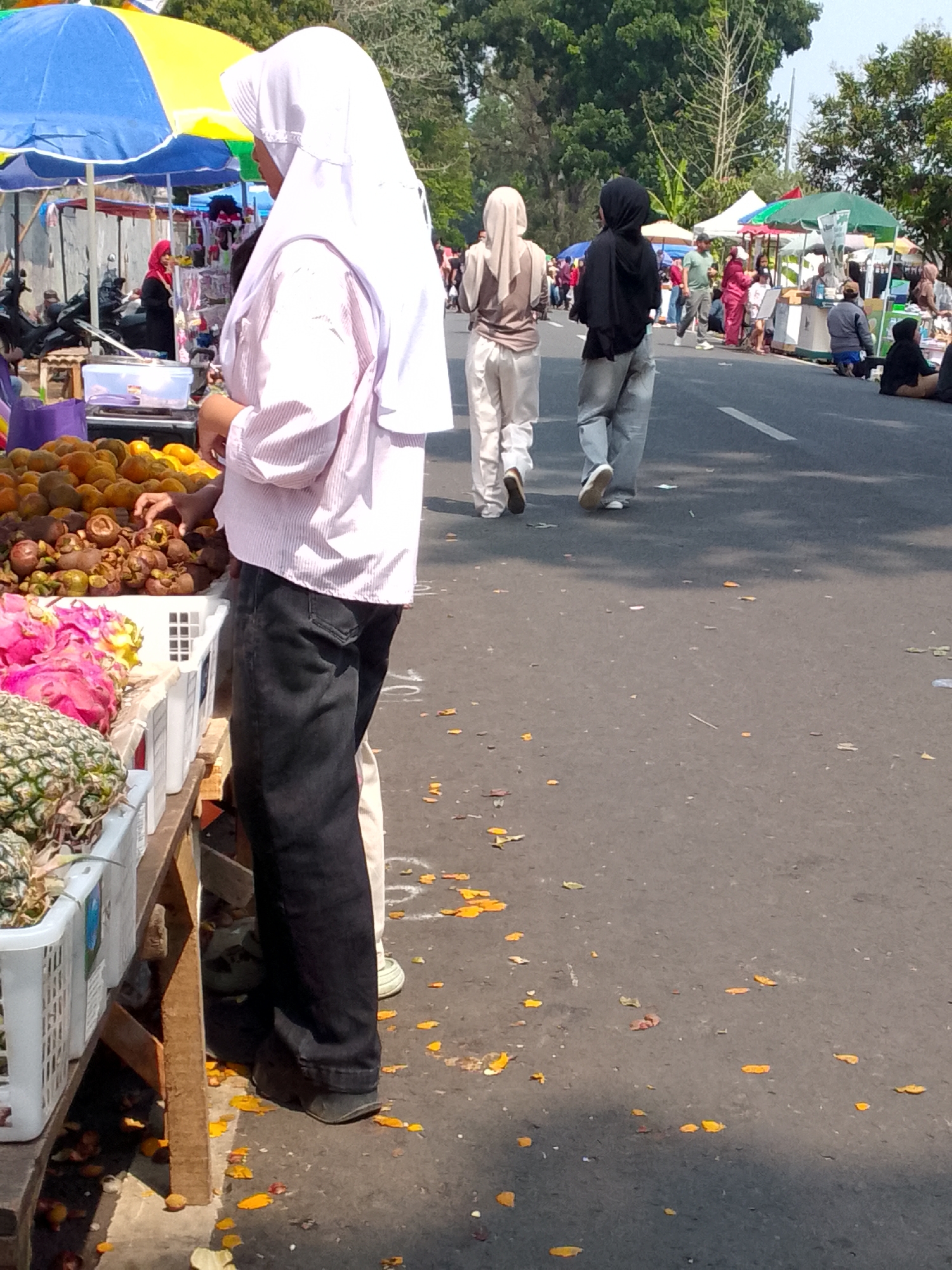 The image depicts an outdoor market scene, likely in Mandalawangi, Indonesia, characterized by stalls set up along a paved road. In the foreground, a person wearing a white hijab and a pink striped shirt is standing near a fruit stall. The stall displays various fruits including pineapples, dragon fruits, oranges, and mangosteens. The person's hands are reaching towards the mangosteens. To the right of the foreground figure, other individuals are visible walking on the road, some wearing hijabs and modest clothing. Several market stalls with colorful umbrellas and displays of goods are set up on the side of the road. The ground is paved and scattered with fallen leaves and petals. The environment suggests a sunny day with trees and foliage visible in the background.