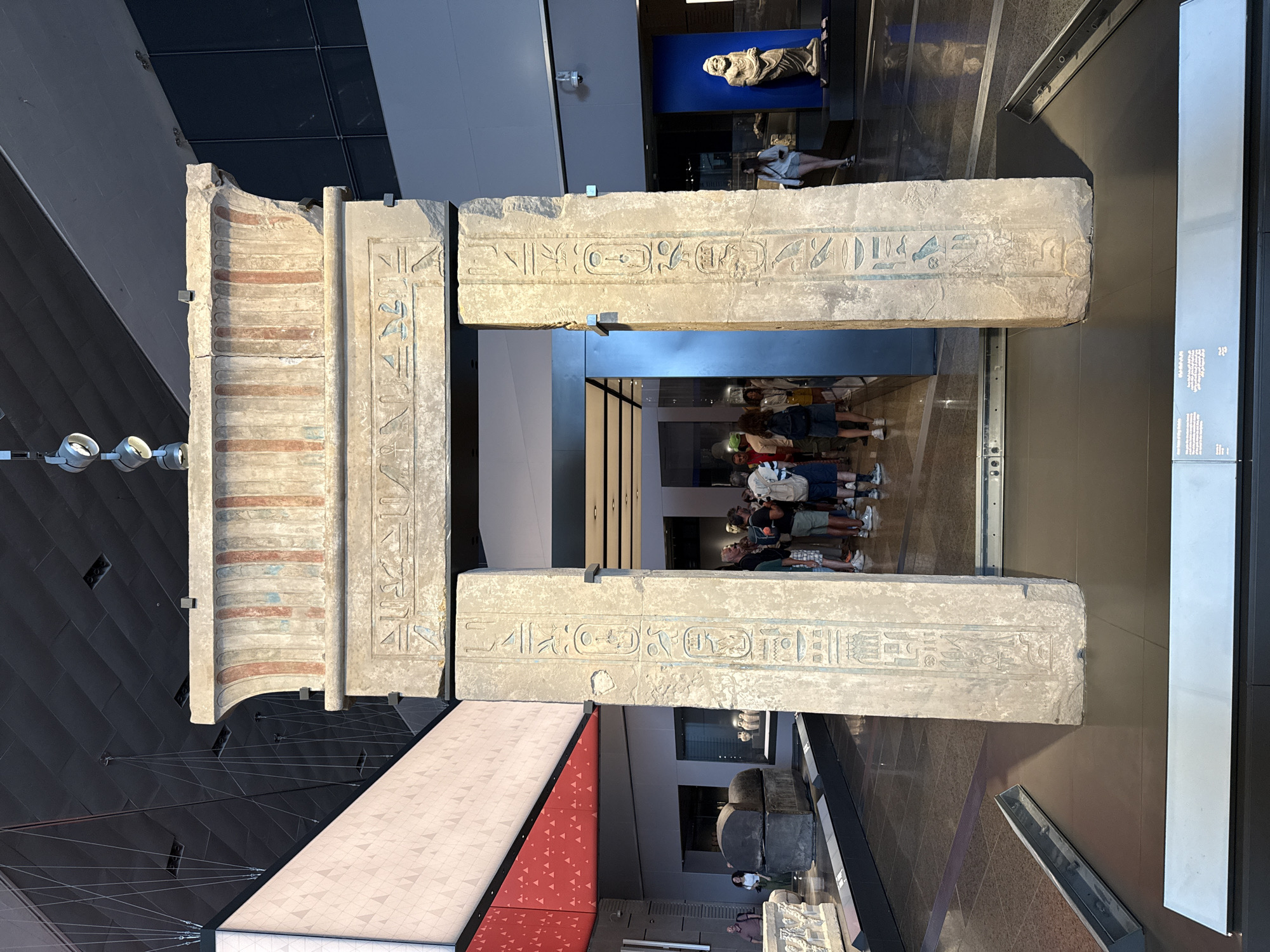 The image shows an ancient Egyptian stone gateway with hieroglyphs carved into it, located in a museum. The gateway is comprised of two upright pillars and a lintel, all made of weathered stone. The lintel features decorative elements and hieroglyphic inscriptions. The pillars also bear hieroglyphic carvings. In the background, several people are visible, some standing and observing the artifact, while others are walking through the museum. The museum environment appears modern, with high ceilings, lighting fixtures, and display cases visible. The context of the city of Kafrat Nassar, Egypt, is not evident from the visual information in the image.