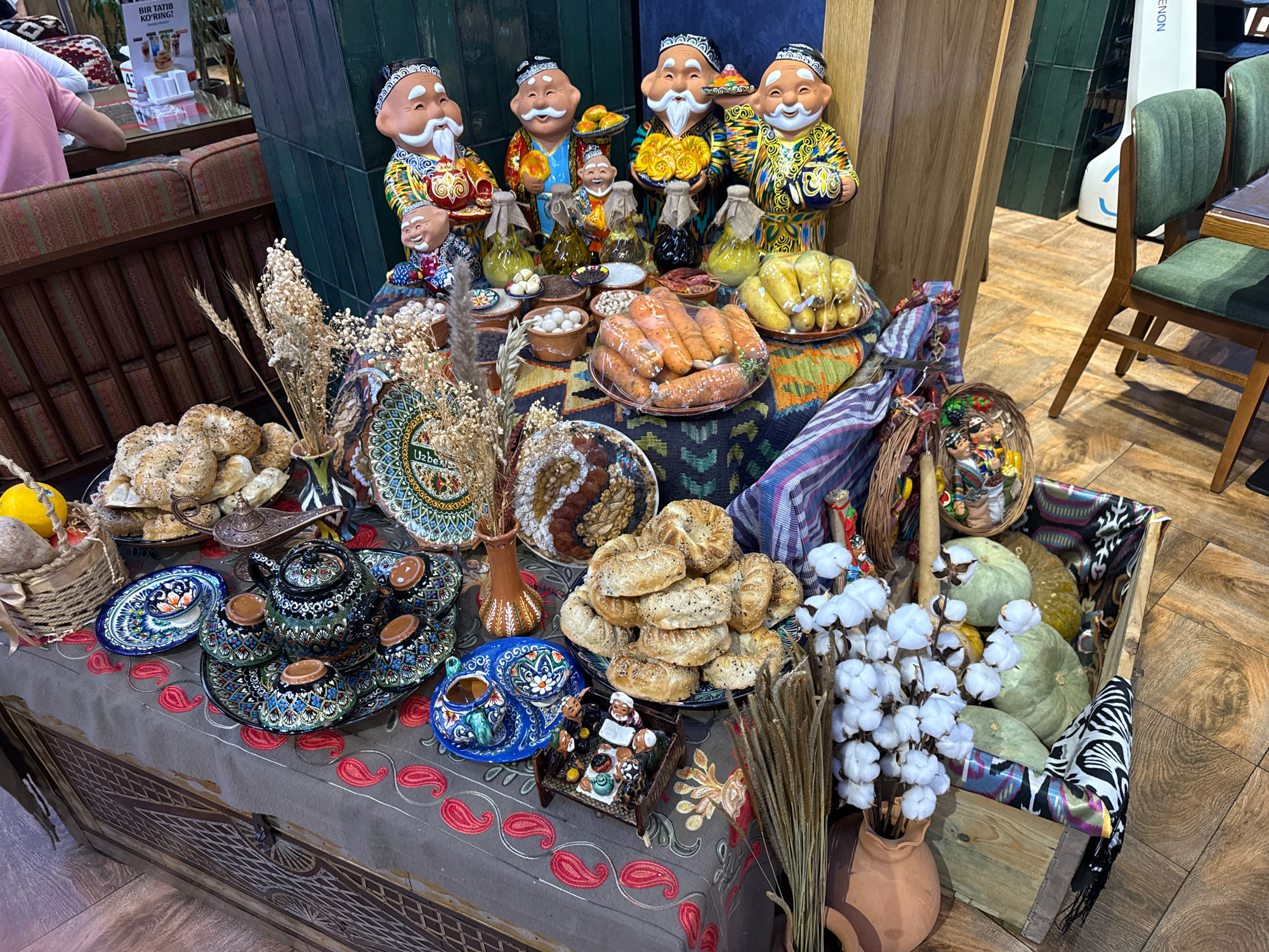 A still life display of traditional Uzbek food and decor is set up on a table covered with a patterned cloth. In the background, several ceramic figures depicting men in traditional clothing are arranged, holding food items and bottles. The foreground features various dishes, including pastries, fruits, vegetables, and a tea set with ornate cups and a teapot. Several vases with dried flowers and cotton bolls are also present. The scene appears to be indoors, possibly in a restaurant or market, with a wooden floor and other furniture visible in the background. The overall impression is one of abundance and cultural celebration.