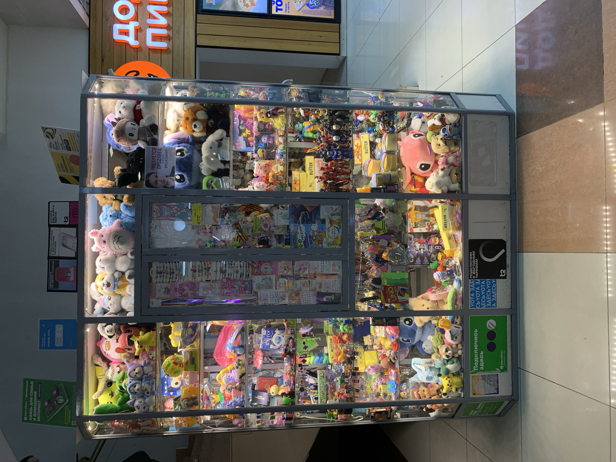 The image depicts a brightly lit, multi-sided display case filled with an assortment of plush toys and action figures. The display case is situated in what appears to be an indoor shopping environment, suggested by the tiled flooring and the presence of commercial signage in the background.
In the center of the display case, several plush toys are prominently featured, including a large blue Stitch character, a pink character resembling a Totoro, and various smaller stuffed animals like dogs and cats. The shelves are densely packed with these items, alongside smaller toys, keychains, and packaging for various goods.
On the exterior of the display case, there are advertisements and informational signs. One sign, in Russian, advertises a telecommunications service, "MegaFon," with an offer for mobile internet. Another section of signage is for "T2," another telecommunications provider.
In the upper right portion of the image, a large, illuminated sign reads "ДОМ ИГРУШЕК" which translates to "TOY HOUSE." This sign is a prominent indicator of the type of establishment. The overall environment suggests a retail space focused on selling toys and related merchandise.
Although the image does not explicitly state Prokopyevsk, Russia, the Russian language signage on the advertisements strongly indicates a location within Russia. The style of the display and the products suggest a typical toy store or a section within a larger department store. No people are visible within the immediate vicinity of the display case. The lighting is artificial and bright, illuminating the contents of the display case. The floor is a combination of light-colored tiles with some darker, possibly granite or marble, sections.