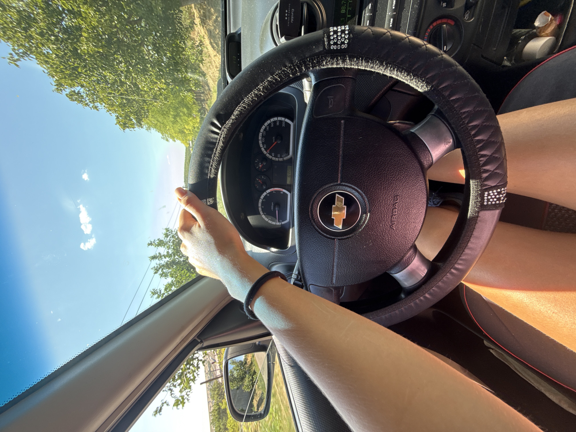 A first-person perspective shot shows a person's arms and legs inside a car. The car's steering wheel is in the foreground, with the driver's left hand resting on it. The steering wheel is covered in a black, quilted material adorned with rhinestones. The dashboard features a Chevrolet emblem. The view through the windshield reveals a bright blue sky with scattered clouds, and green trees line a rural road, suggesting the location is outside of a city. The road and surrounding landscape indicate a countryside setting, possibly in or around Basarabeasca, Moldova, given the rural scenery. The sun is shining brightly, casting shadows inside the car.